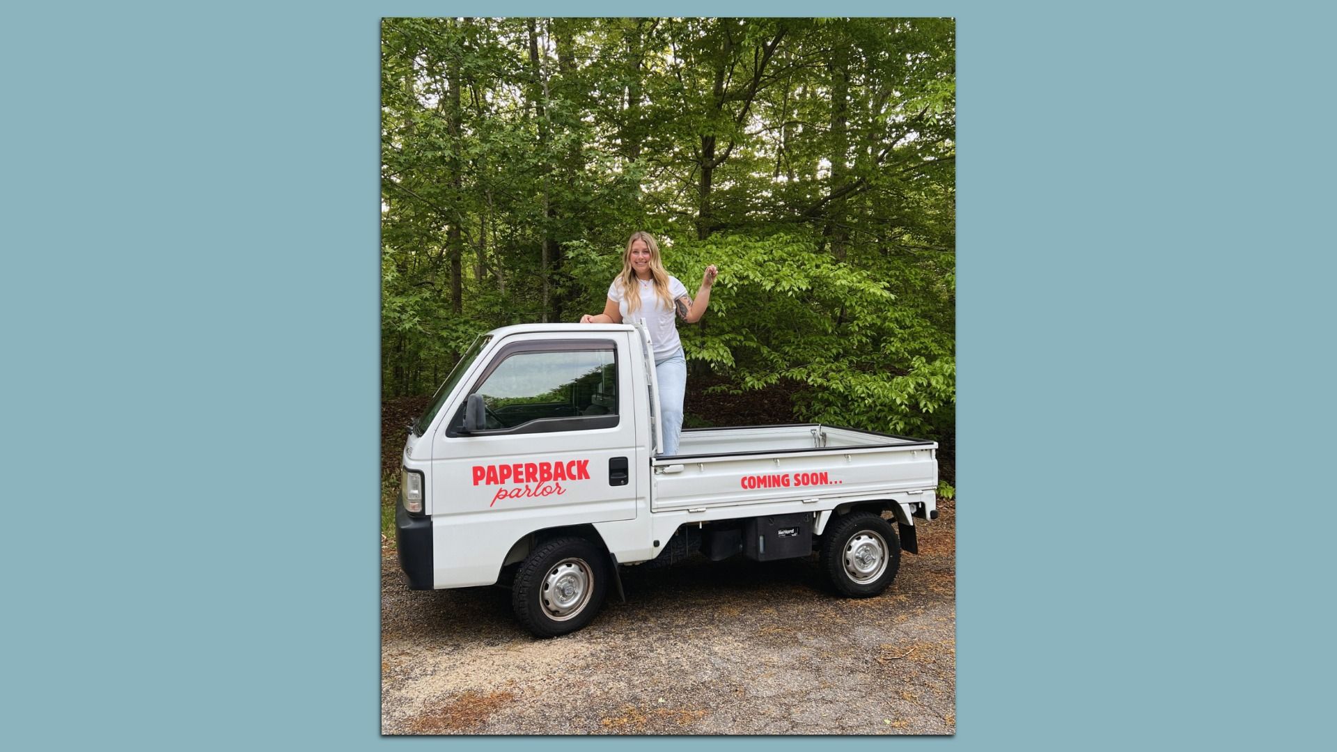 Smiling woman stands in the open bed of a white pickup truck, logo "PAPERBACK parlor" on the side; "COMING SOON..." on the tailgate, with dense green trees in the background.