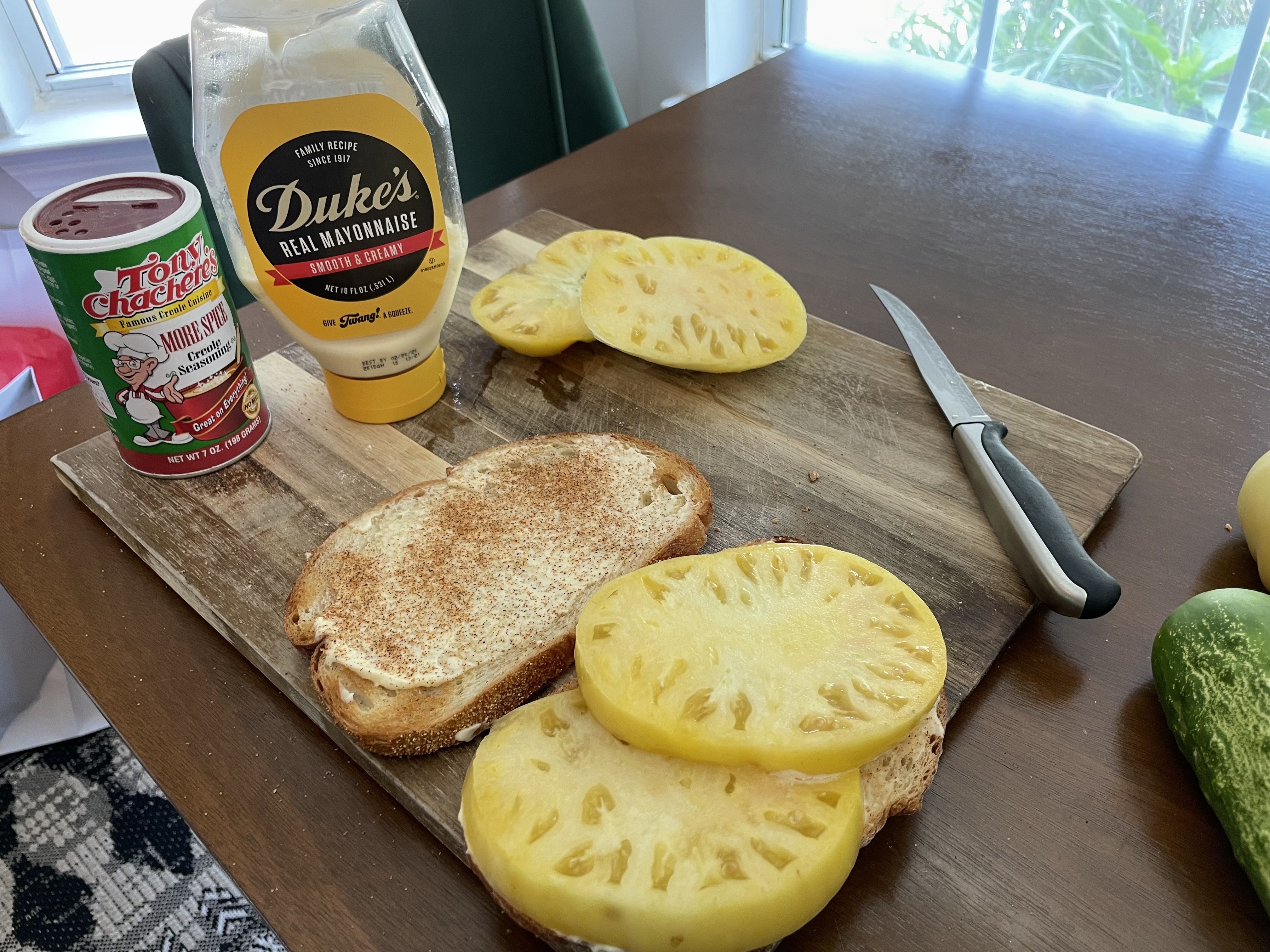 A tomato sandwich-in-progress is shown on a cutting board on a table, with a bottle of Duke's Mayonnaise and Tony Chachere's creole seasoning .