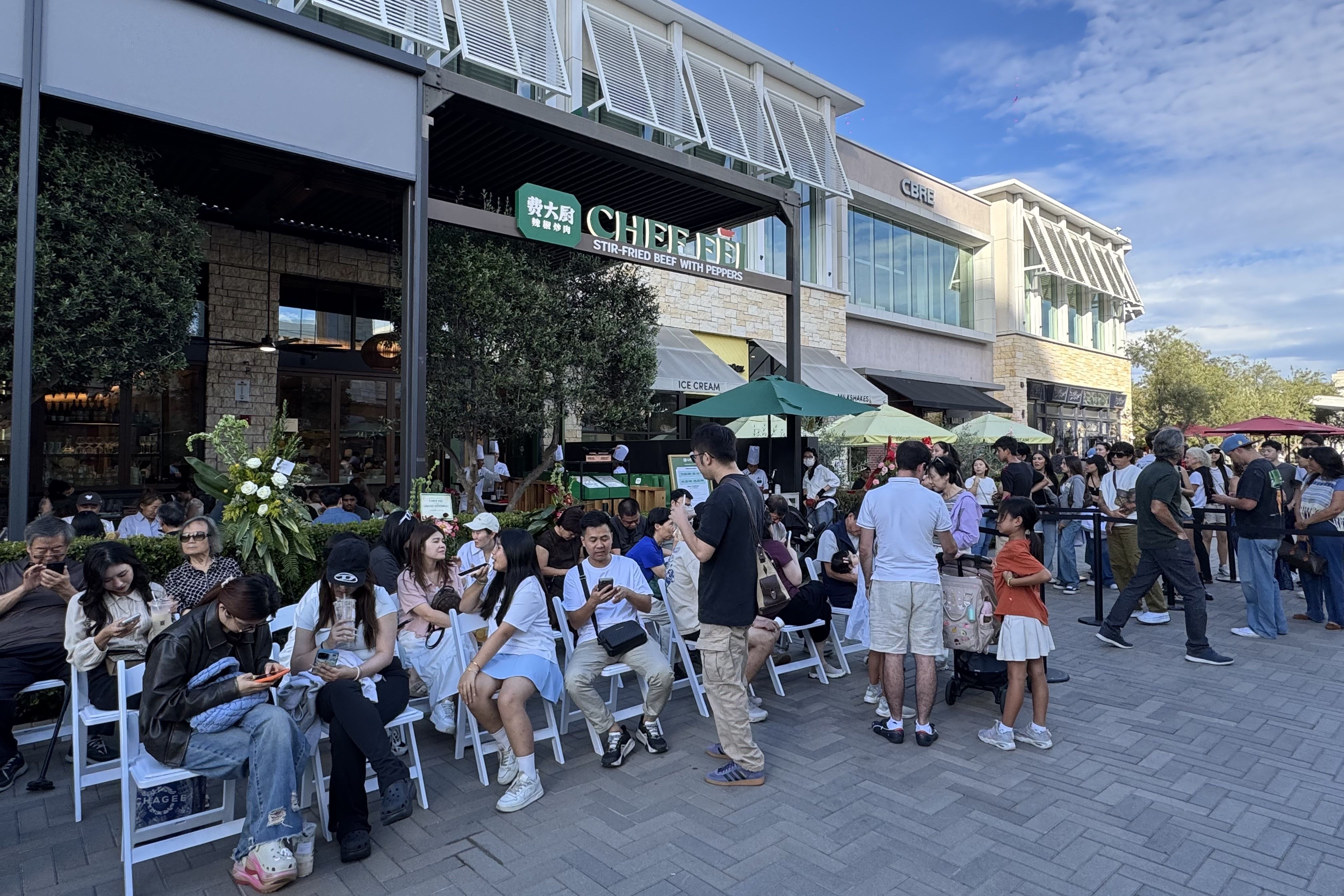 Busy outdoor dining area at a shopping plaza. A restaurant sign reads "CHEF" and "Stir-Fried Beef with Peppers." People sit on white chairs under green umbrellas, with glass storefronts and a blue sky.