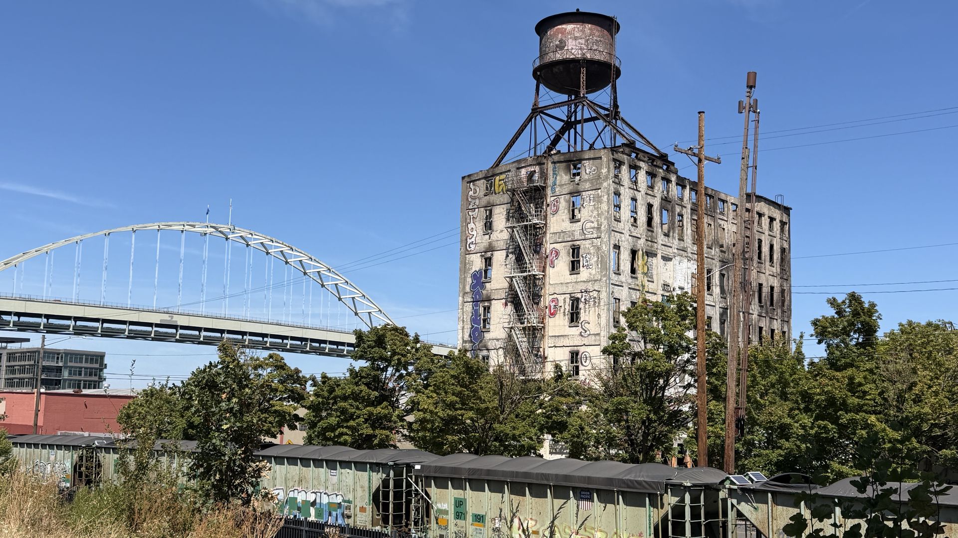 A dilapidated brick factory with a large rusted water tower on a metal frame, graffiti-covered walls, and freight train cars in front; a white arched bridge and blue sky complete the scene.