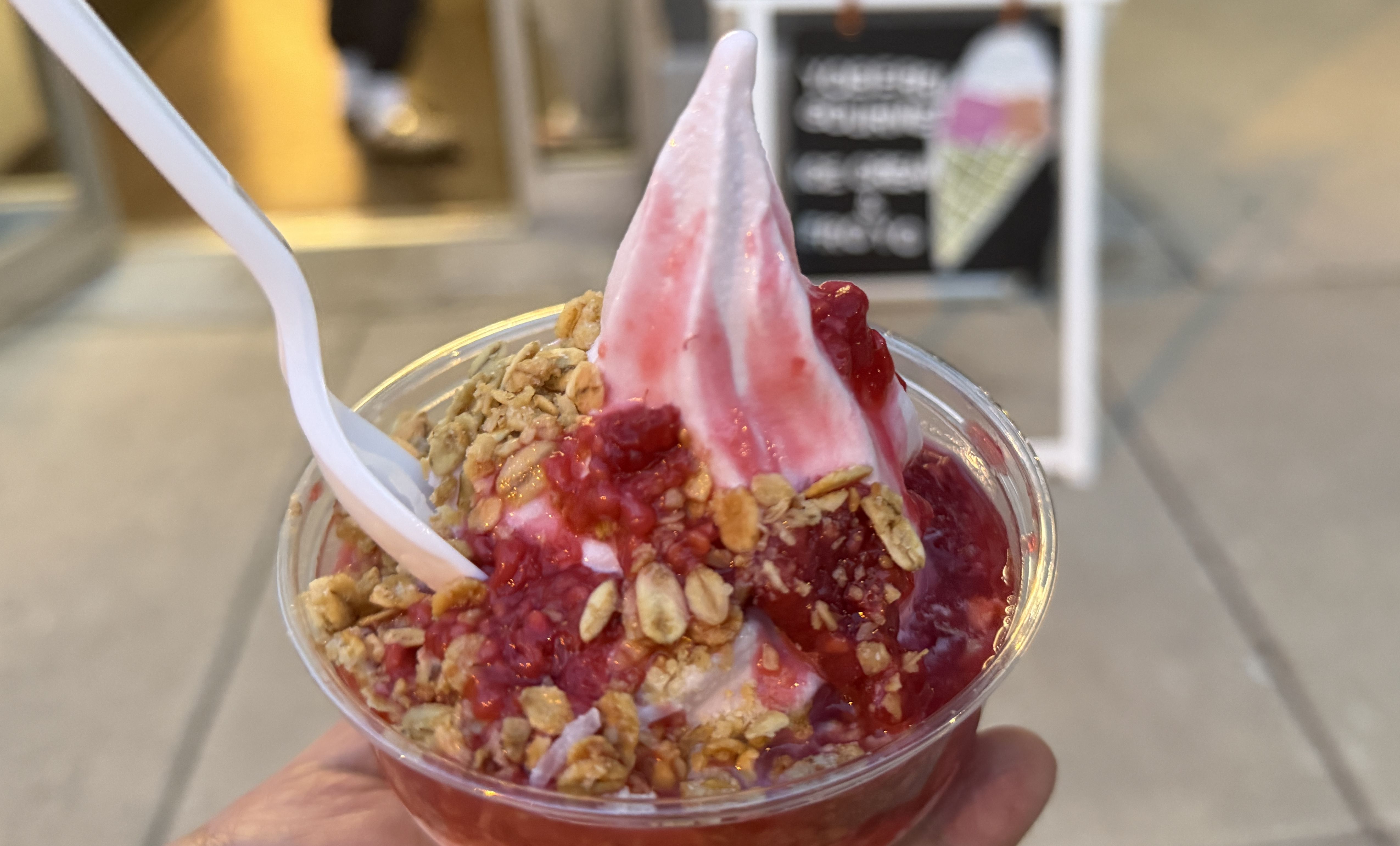 Close-up of a dessert cup with pink swirled soft-serve, red berry sauce, and granola topping; a white straw sticks out, with a blurred cafe interior in the background.