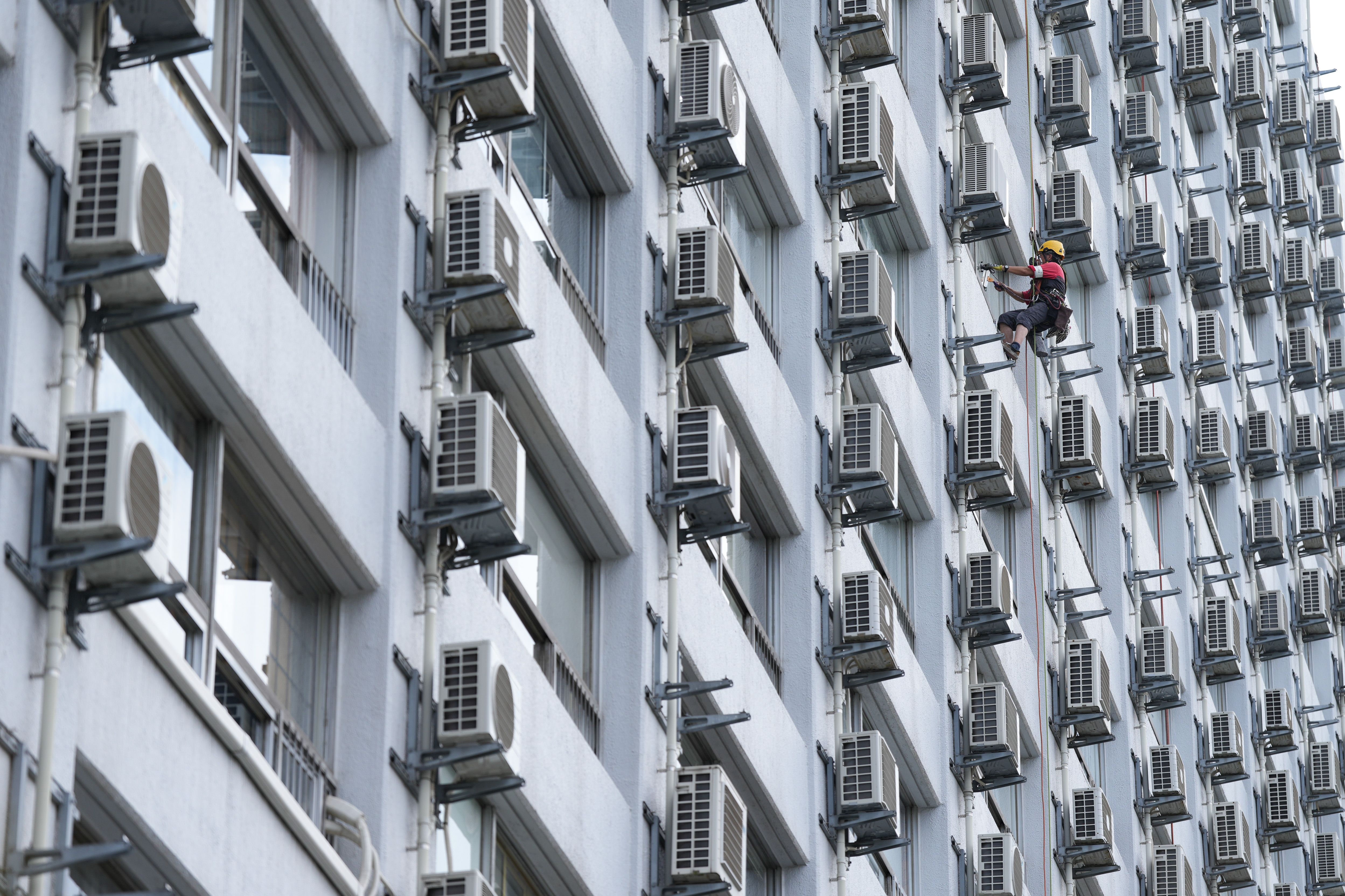 A photograph showing the side of a building with numerous air conditioning units. A single worker is seen amongs the units scaling the wall. 