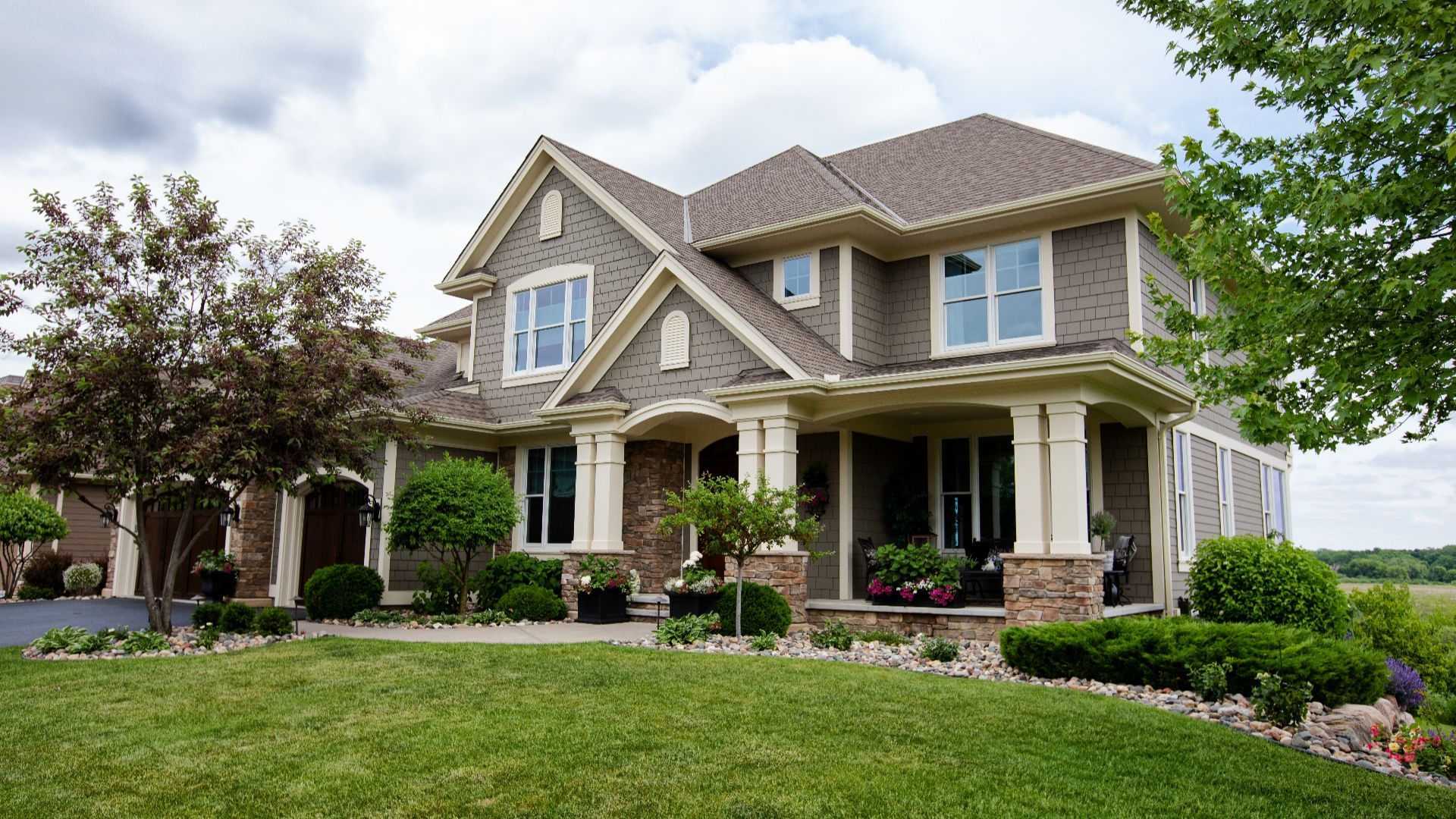 Two-story suburban home with gray siding and stone accents, a wide front porch supported by columns, manicured lawn, and trees framing the house under a partly cloudy sky.