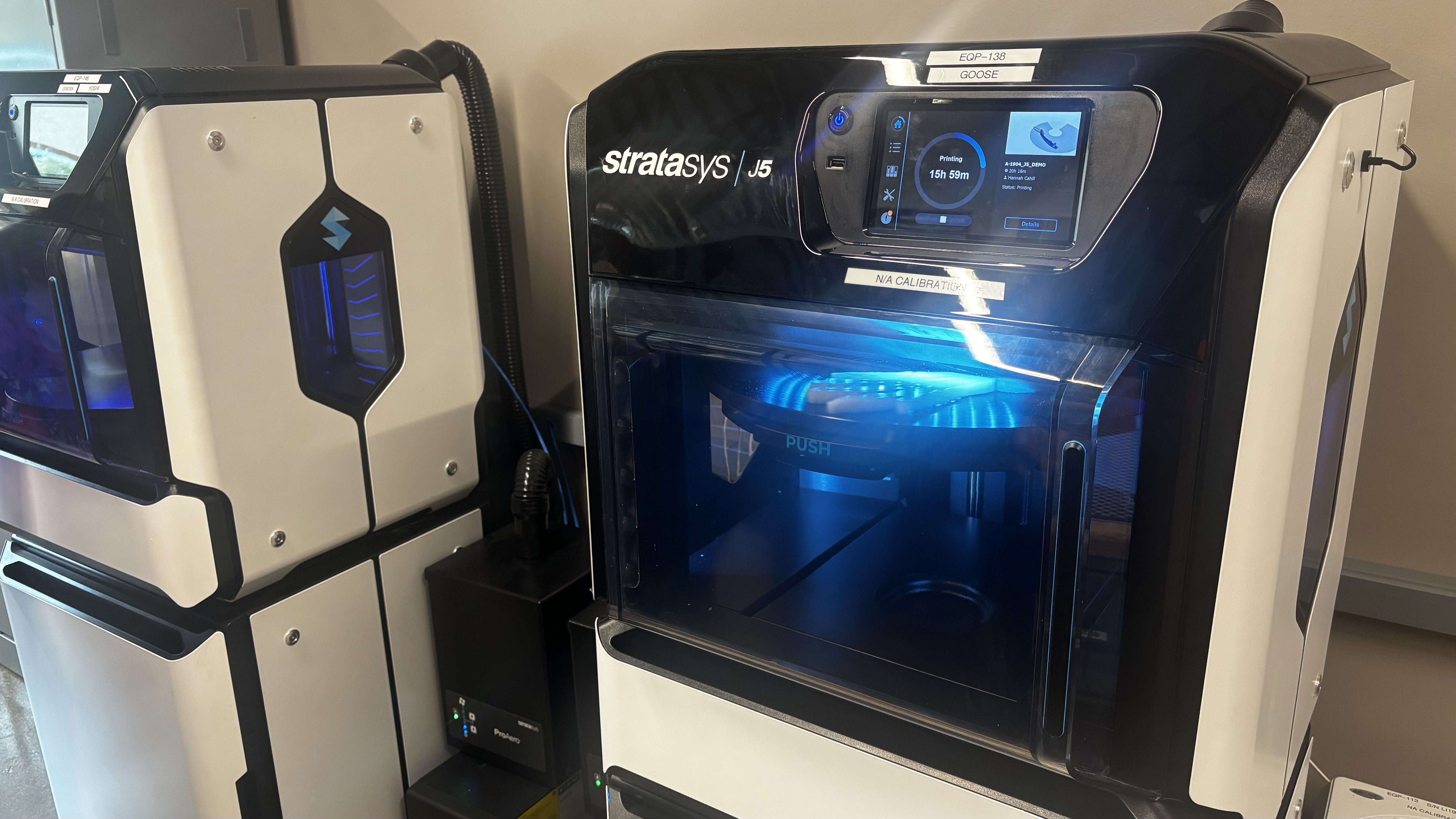Row of Stratasys 3D printers in a lab: the right unit has a blue-lit interior and touchscreen, labeled Stratasys J5; a second machine with a windowed door sits beside it.