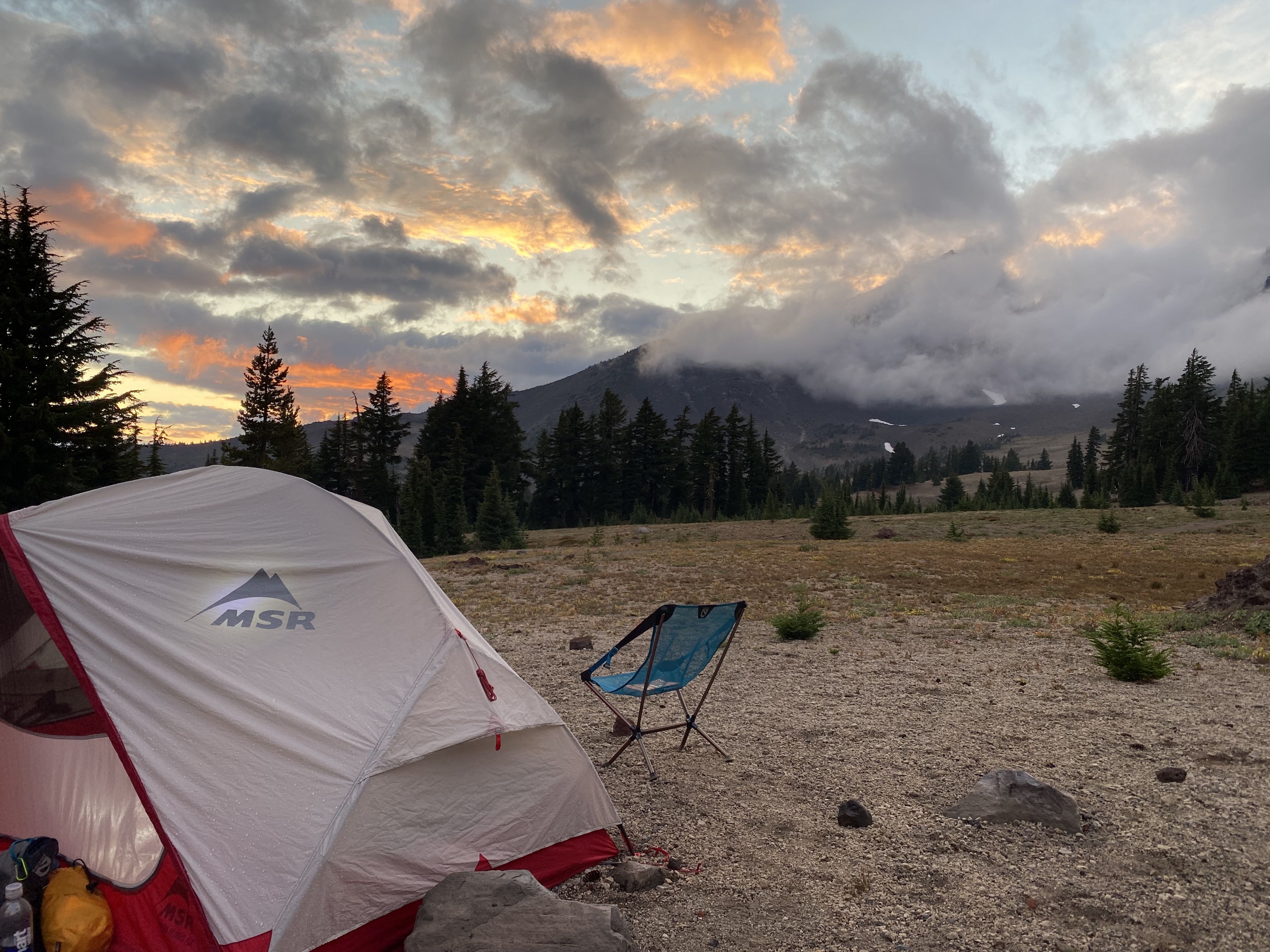 White and red MSR tent and blue camping chair on rocky ground at sunset with pine trees and fog-covered mountains in the background under a cloudy sky with orange and purple hues.