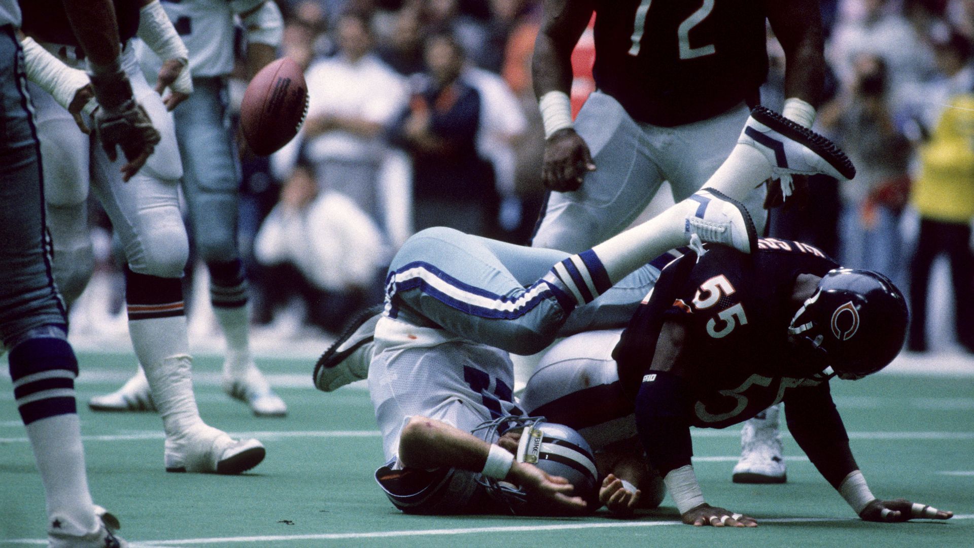 A football player in white and blue falls backwards while a player in black and orange, number 55, tackles him on a green field during a game with blurred audience in the background.