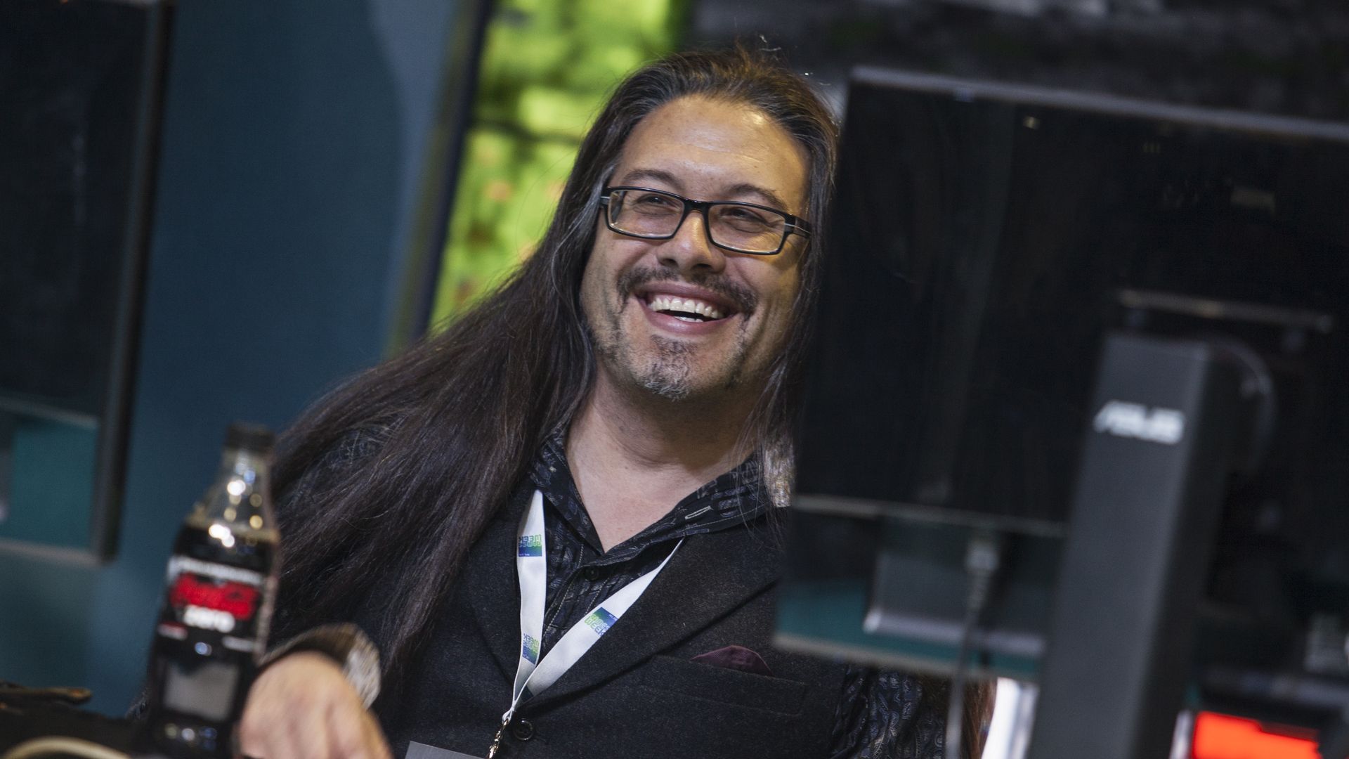A man with elbow-length hair sits at a computer, smiling