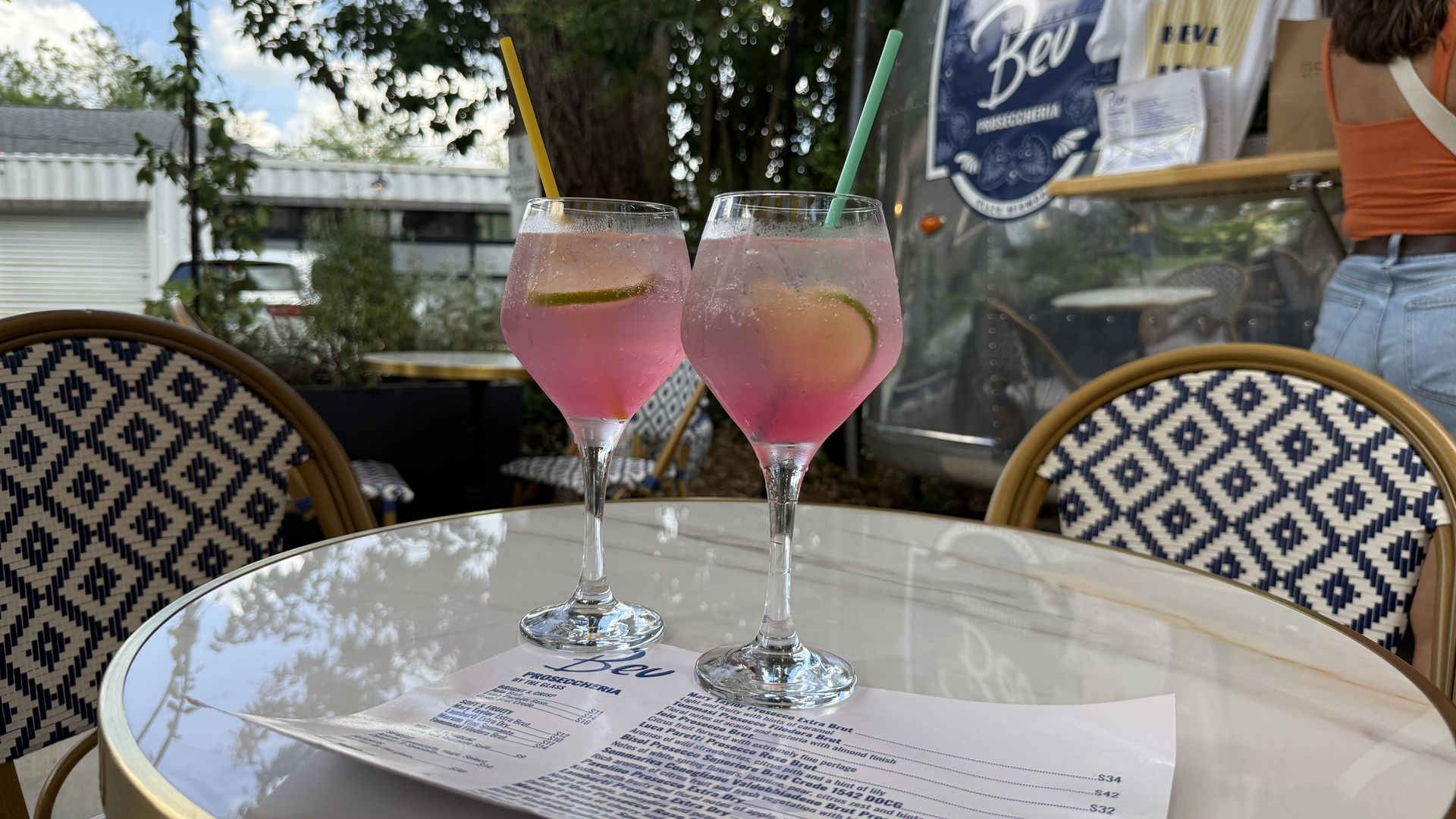 Two pink cocktails with lime wedges and colorful straws sit on a glass patio table. Wicker chairs with a blue-and-white pattern surround the scene, with a shiny food trailer and a person in orange in the background.