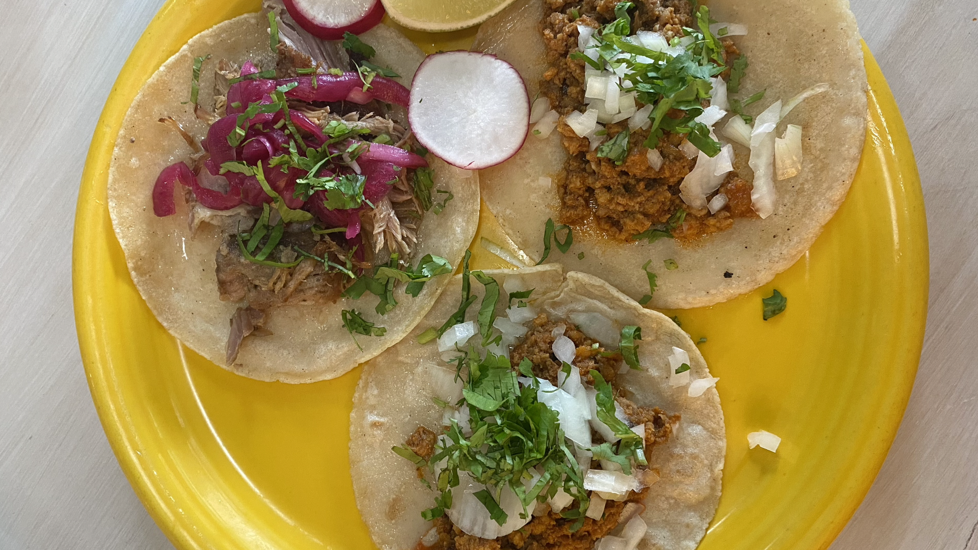 Three open-face tacos on a bright yellow plate with a lime wedge and radish on the side.