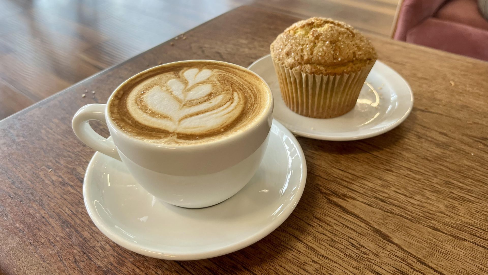 A latte with art and a muffin on white dishware on a wooden coffee table.