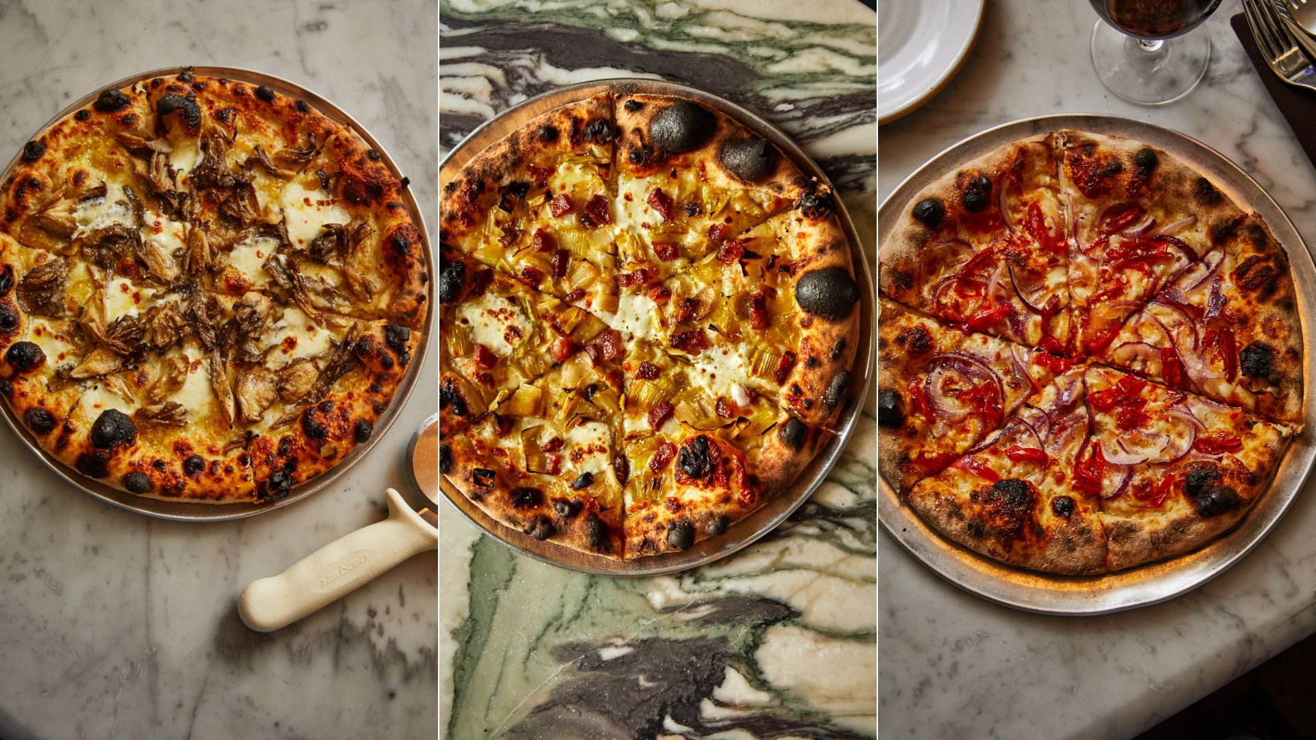 Three wood-fired pizzas on metal trays on marble tables: one with mushrooms and cheese, one with pineapple and bacon, one with red onions and red peppers, all with charred crusts.