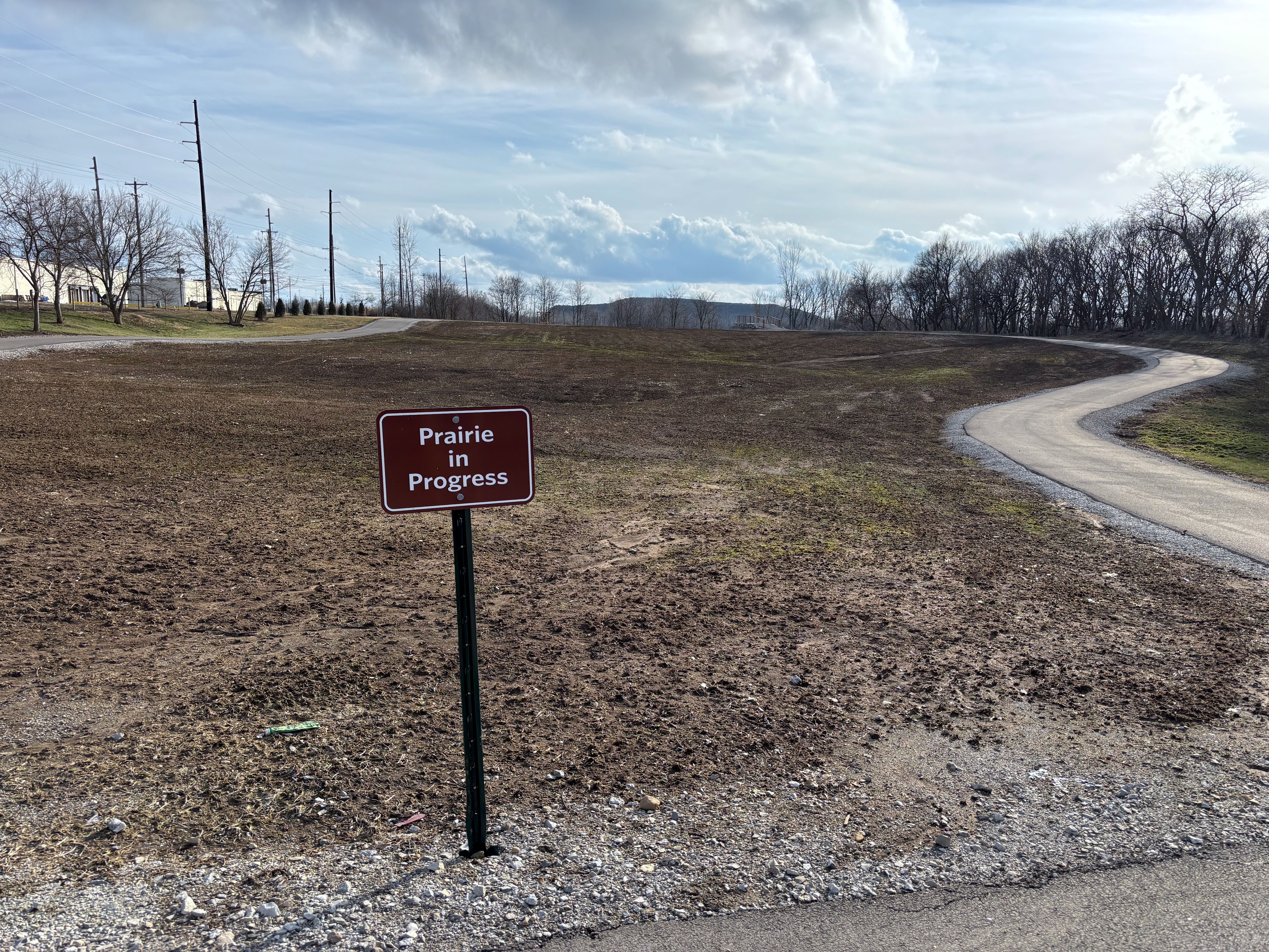 Brown field with sparse grass under a partly cloudy sky, a red sign saying "Prairie in Progress," and a winding paved path on the right, bordered by bare trees and power lines in the distance.