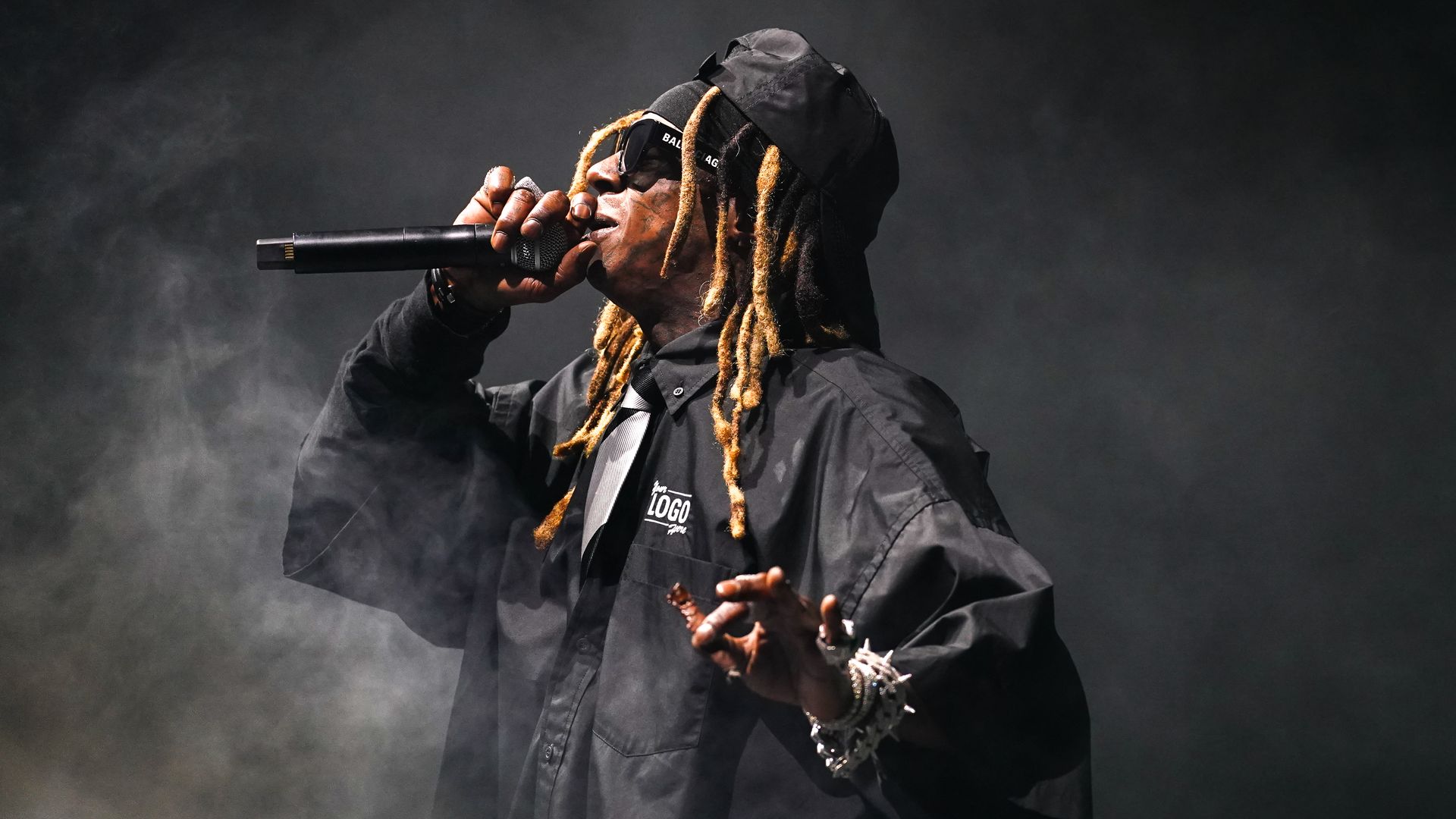 Lil Wayne performs with blond dreadlocks, wearing a black hoodie and sunglasses, singing into a microphone on a smoky, dimly lit stage.