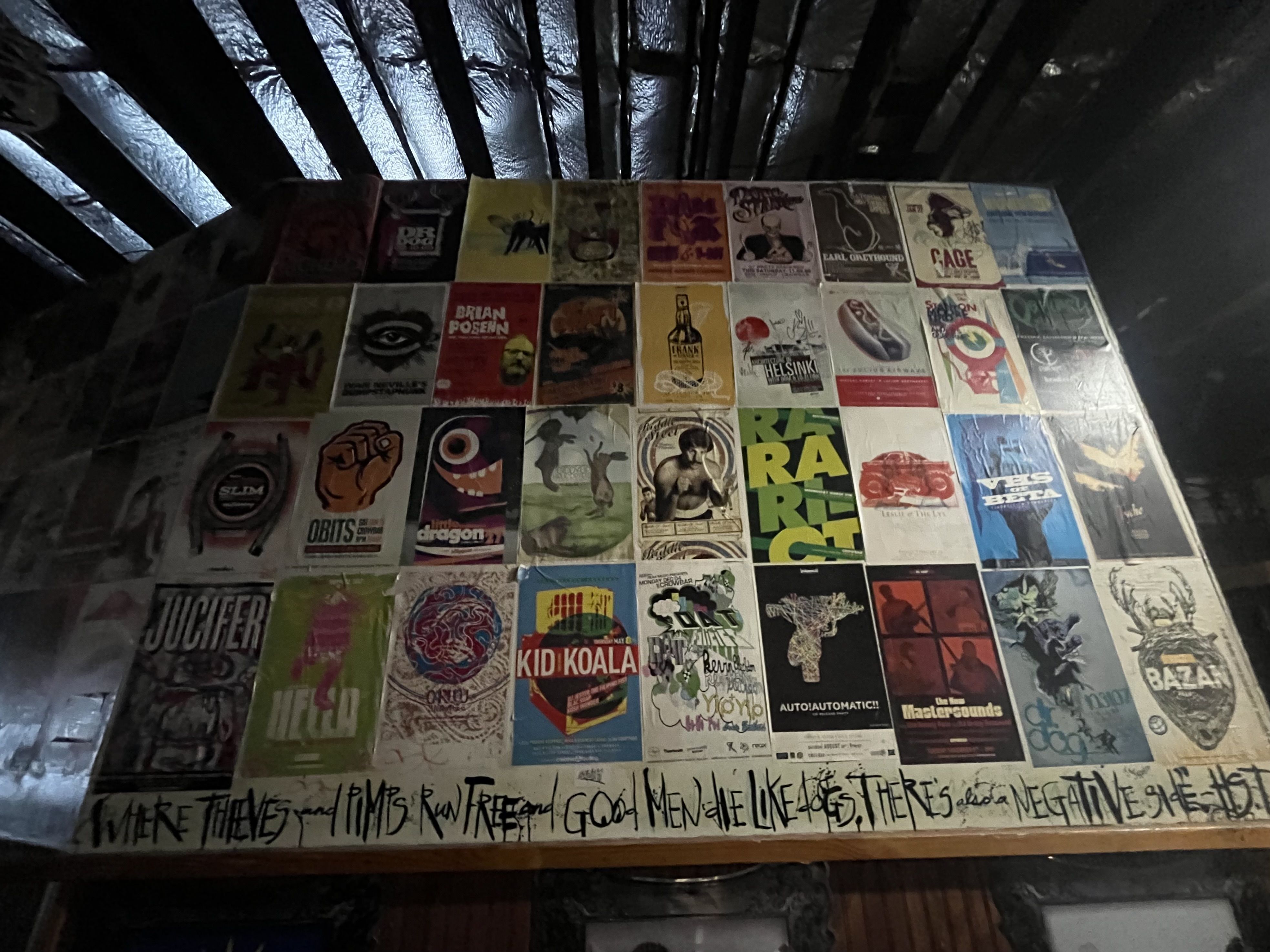 Dim room with a wall covered in colorful posters arranged in a grid, under an exposed slanted ceiling. Graffiti runs along the bottom; posters feature varied art and text like "KID KOALA".