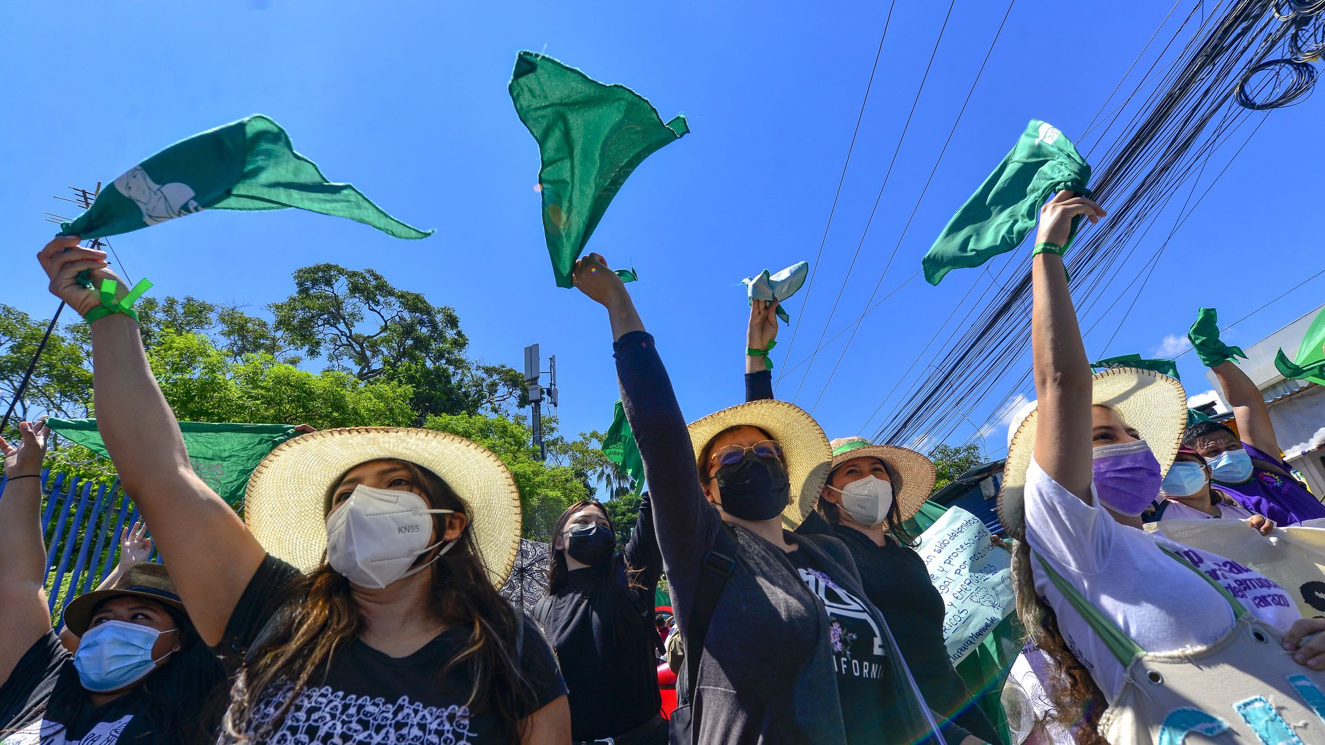 Women in El Salvador wave green flags while protesting for abortion rights outside against the backdrop of a bright blue sky. 