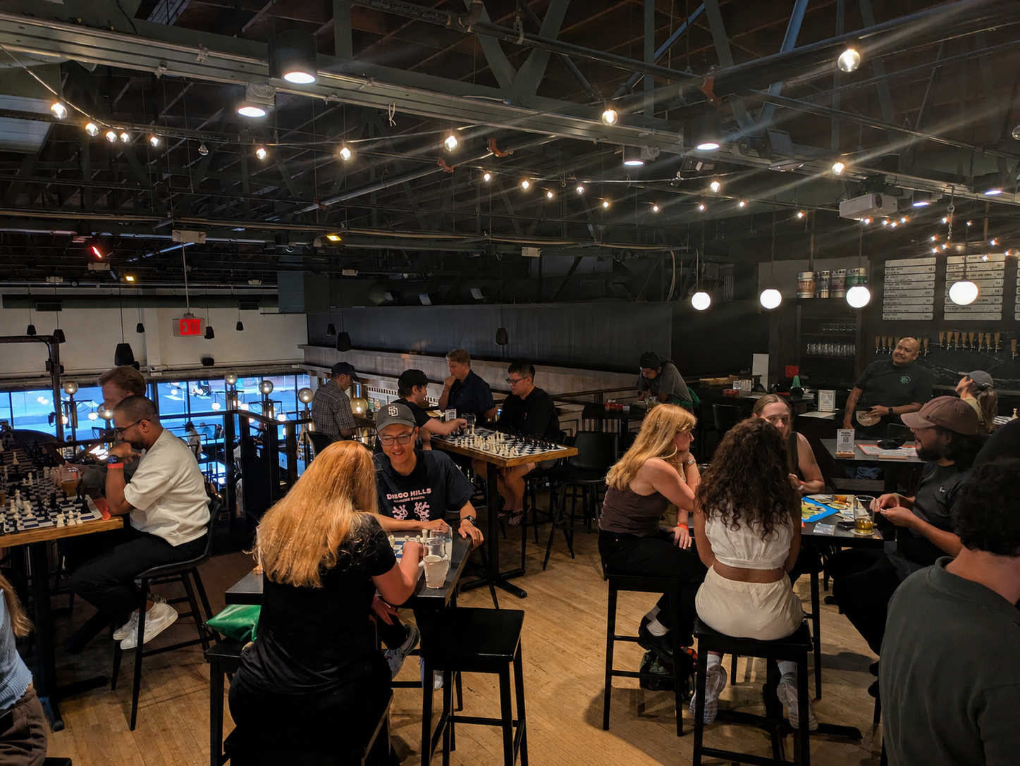 Cafe scene with an exposed ceiling and string lights; groups play chess at tall tables, bar area visible in back-right, warm ambience.