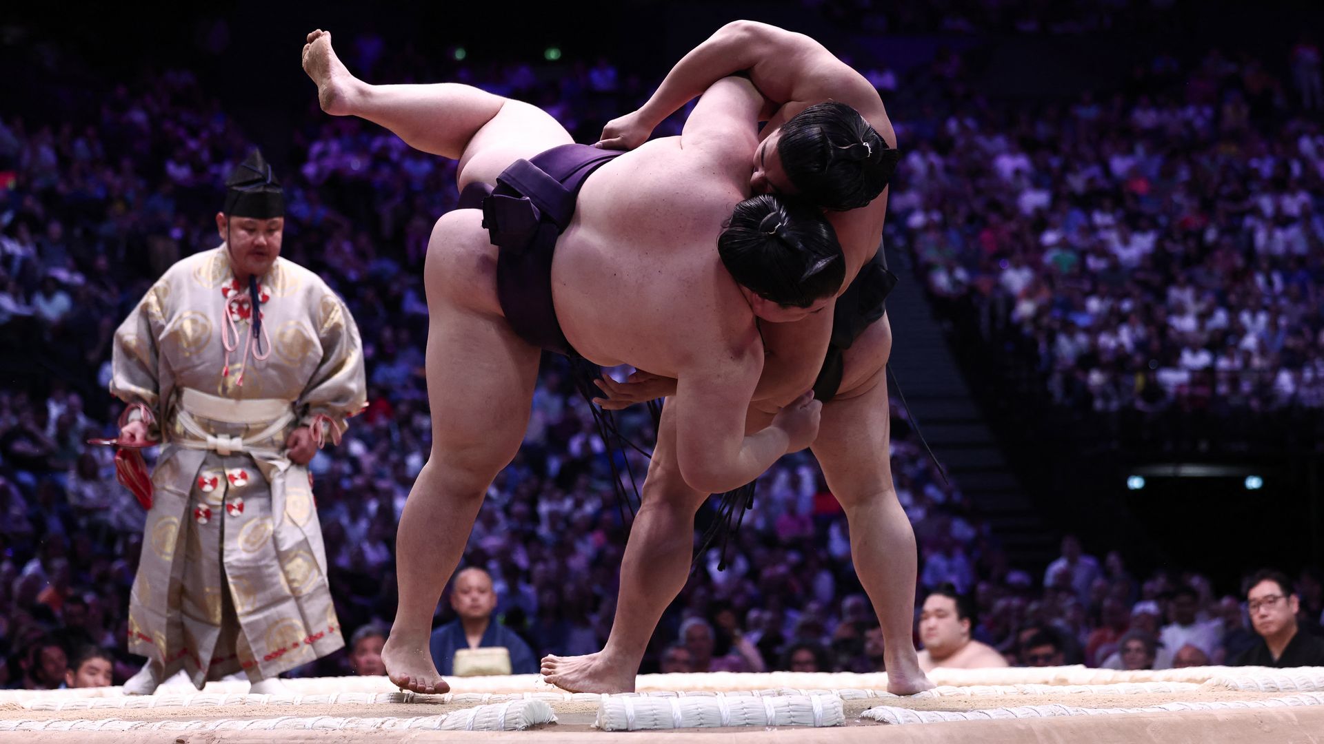 A sumo wrestlers throws another on to the ground in a clay ring while a referee looks on. The wrestling match is taking place in a darkened auditorium with a large crowd watching.