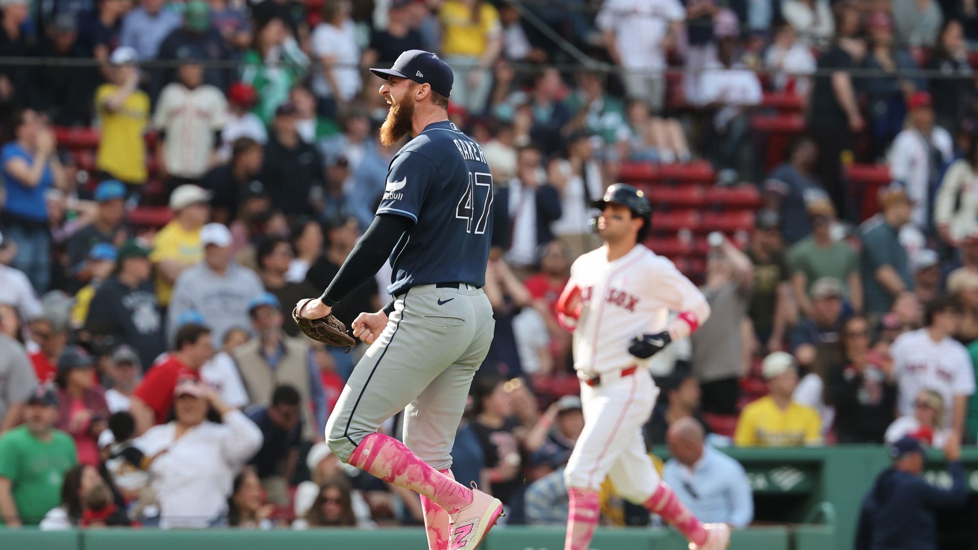 A pitcher wearing a dark Blue Rays uniform and pink socks celebrates after securing the winning out, as a Red Sox player rounds the bases behind him.