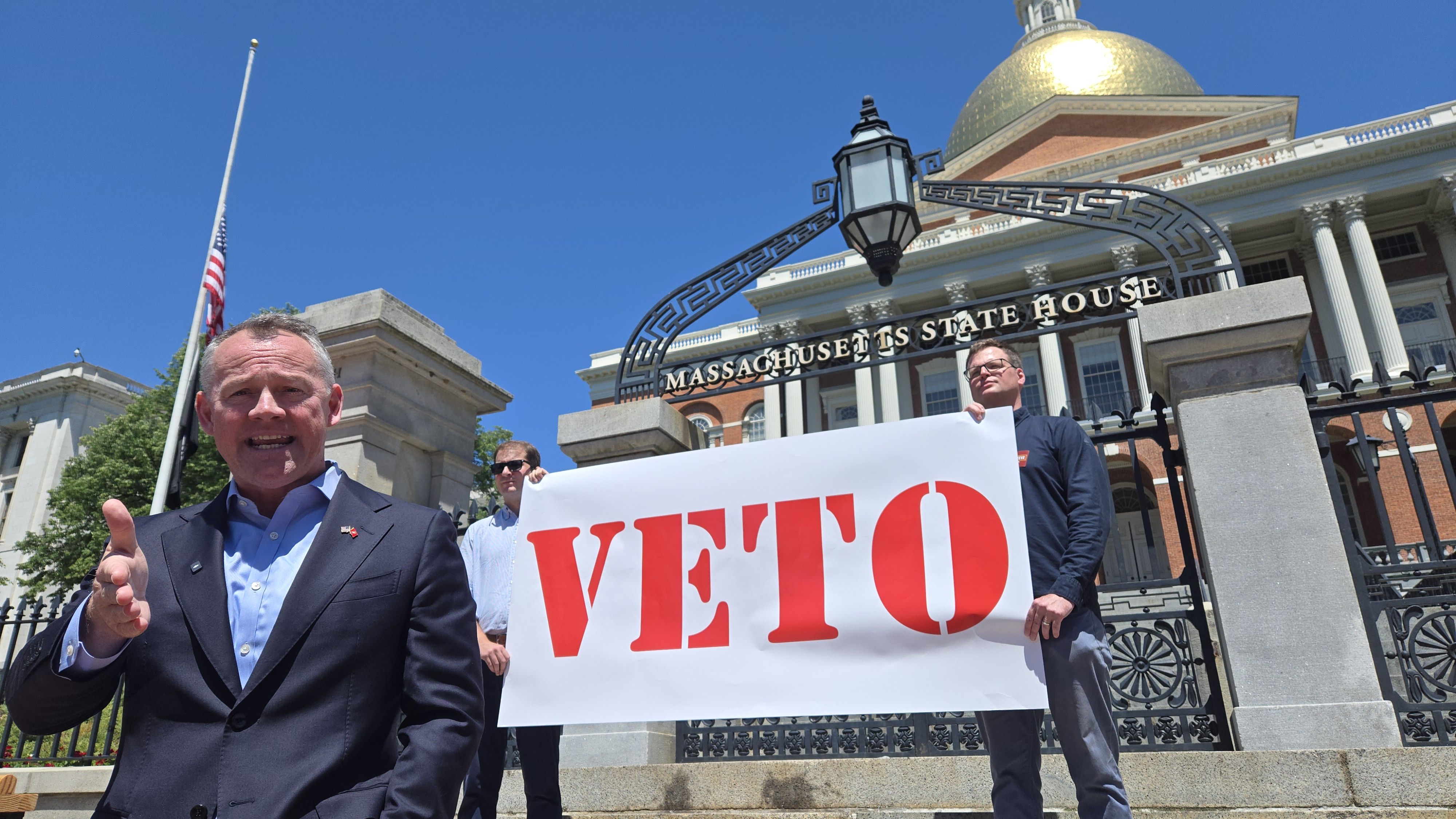 A man in a dark suit gives a thumbs-up while holding a large white sign reading "VETO" in bold red letters, in front of the Massachusetts State House with its gold dome against a bright blue sky.