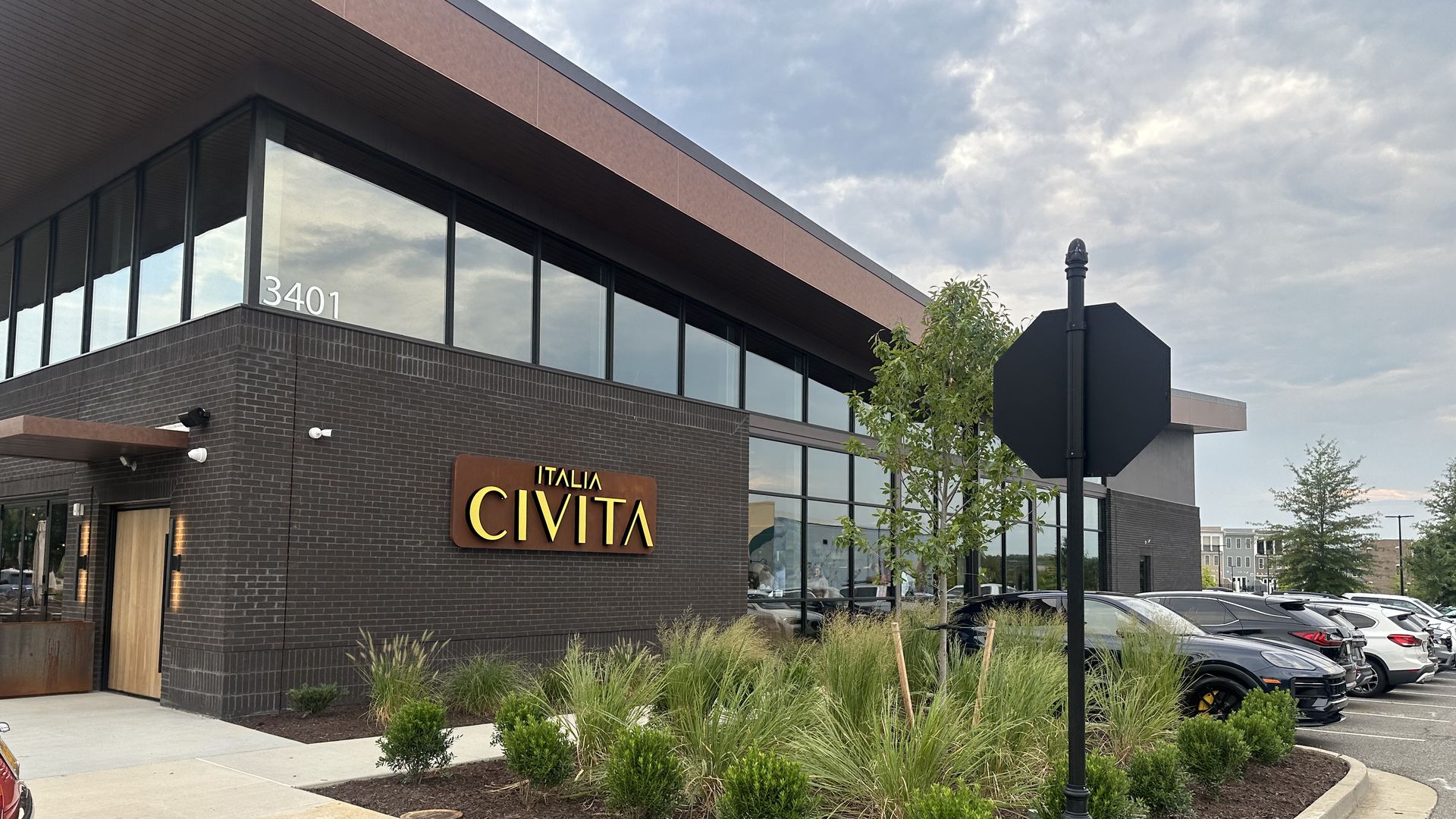 Exterior view of a modern brick building with large glass windows and a brown sign reading "Italia Civita" at 3401. A parking lot, landscaped shrubs, a black street lamp, and a cloudy blue sky.