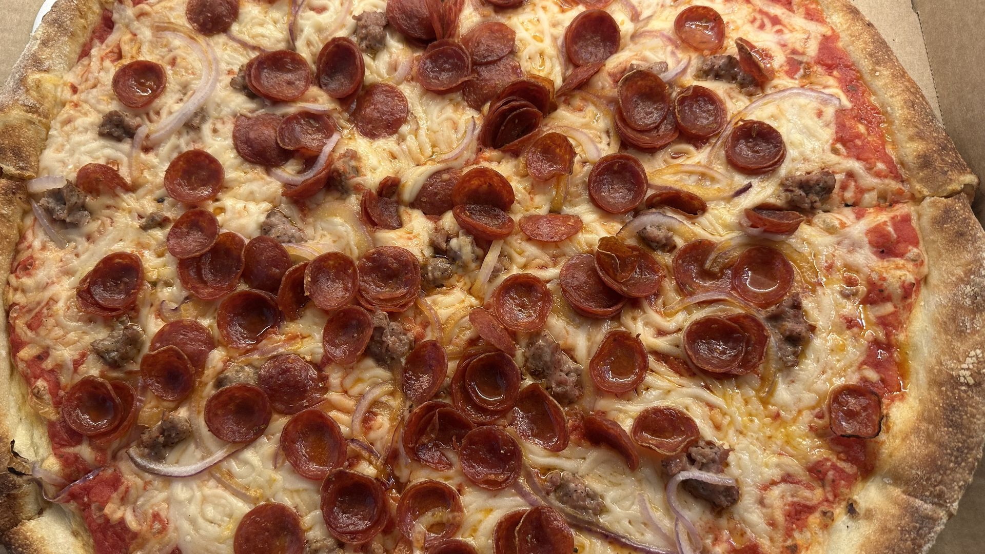 A large pepperoni pizza in a cardboard box, topped with pepperoni slices, onions, sausage, and bubbly melted cheese over tomato sauce and a golden crust.