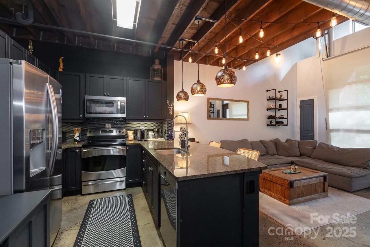 Modern kitchen and living area with black cabinets, stainless steel appliances, granite island, hanging pendant lights, gray sectional sofa, wooden coffee table, high wood ceiling with string lights.