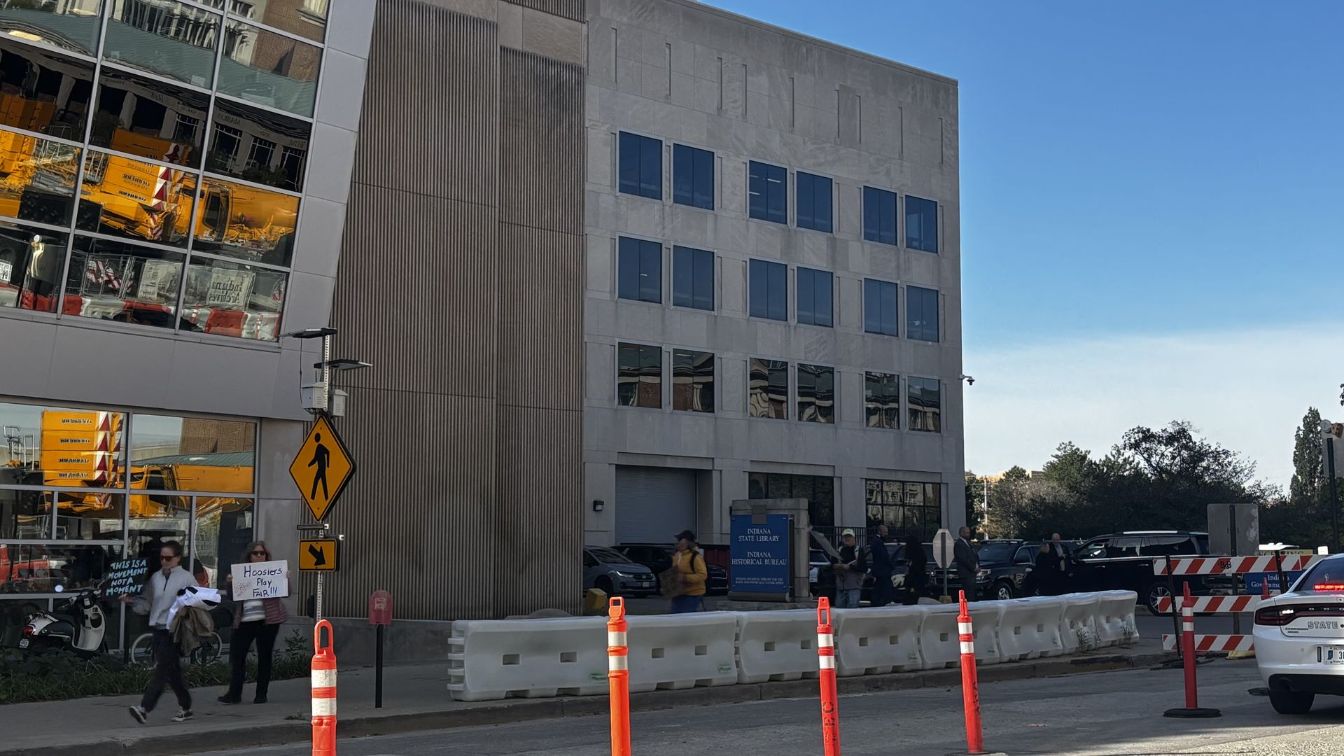 Sidewalk in front of Indiana State Library with people holding protest signs, orange traffic poles, and barriers under a clear blue sky.