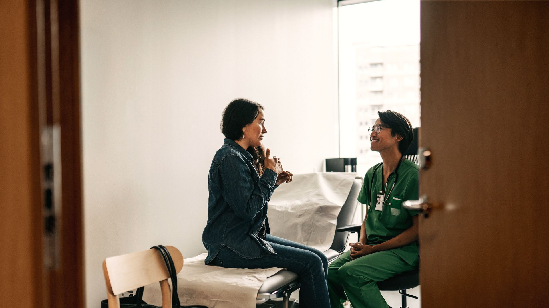 A healthcare worker in green scrubs sits on a chair opposite a patient on an examination bed, in a bright clinic room with a window behind them.