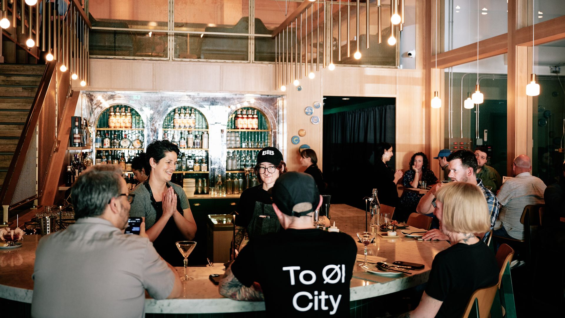 People gather and chat around a marble bar inside Kachka Fabrika, a cocktail bar in Portland, Oregon. Bottles and glassware are displayed on backlit shelves, with modern pendant lights overhead. 