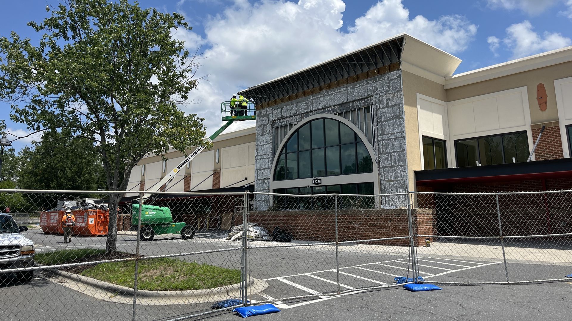 Construction scene at a storefront: two workers in yellow vests on a green lift atop a stone facade, large arched window, beige walls, chain-link fence, and a tree against a blue sky with clouds.