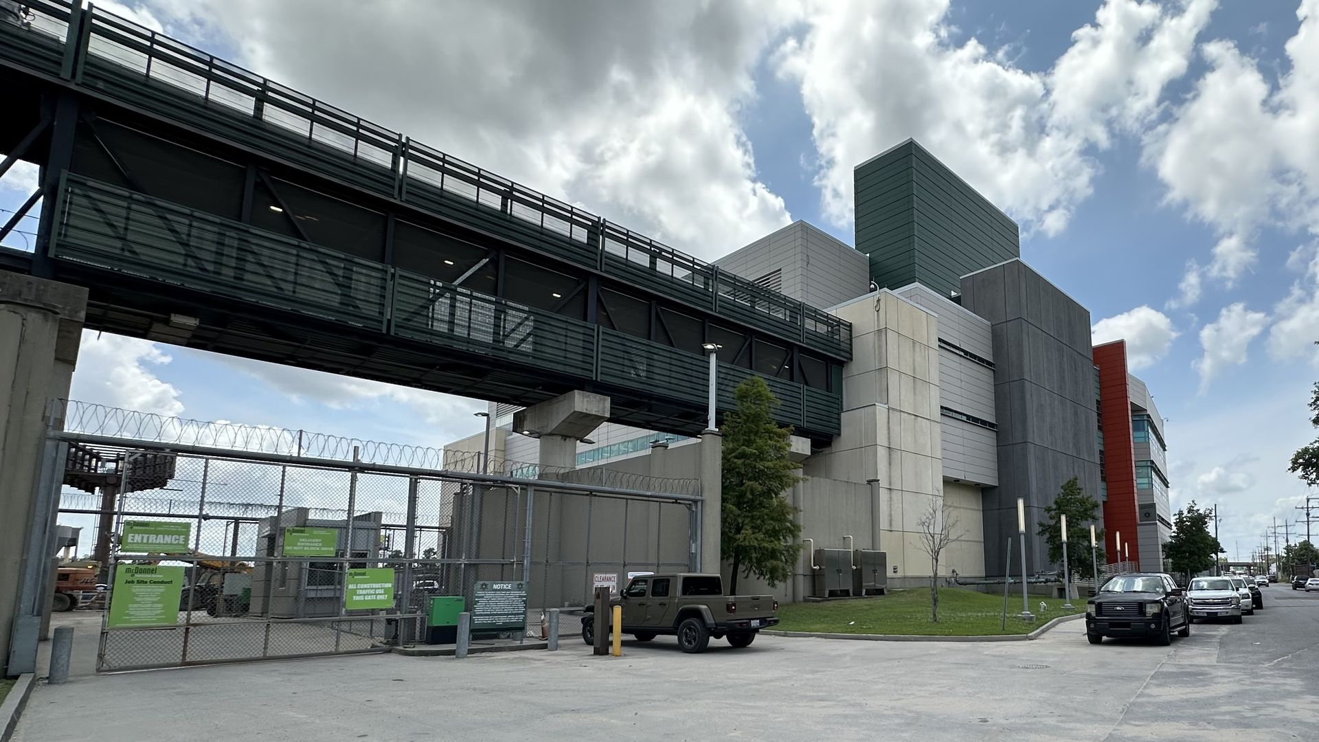 Modern industrial building with multiple colored sections, a covered overhead walkway, fenced construction area, parked cars along the street, and a partly cloudy blue sky.