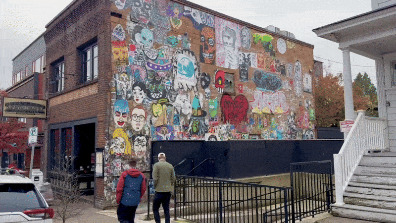 A moving image shows two people walking down the street looking to their right to see a building with colorful street art on the side brick wall.