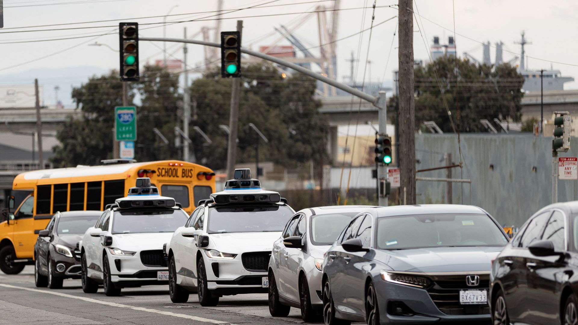 Cars and a yellow school bus stopped at a green traffic light on an urban street. Two white cars with roof-mounted sensors suggest autonomous vehicles in the lineup.