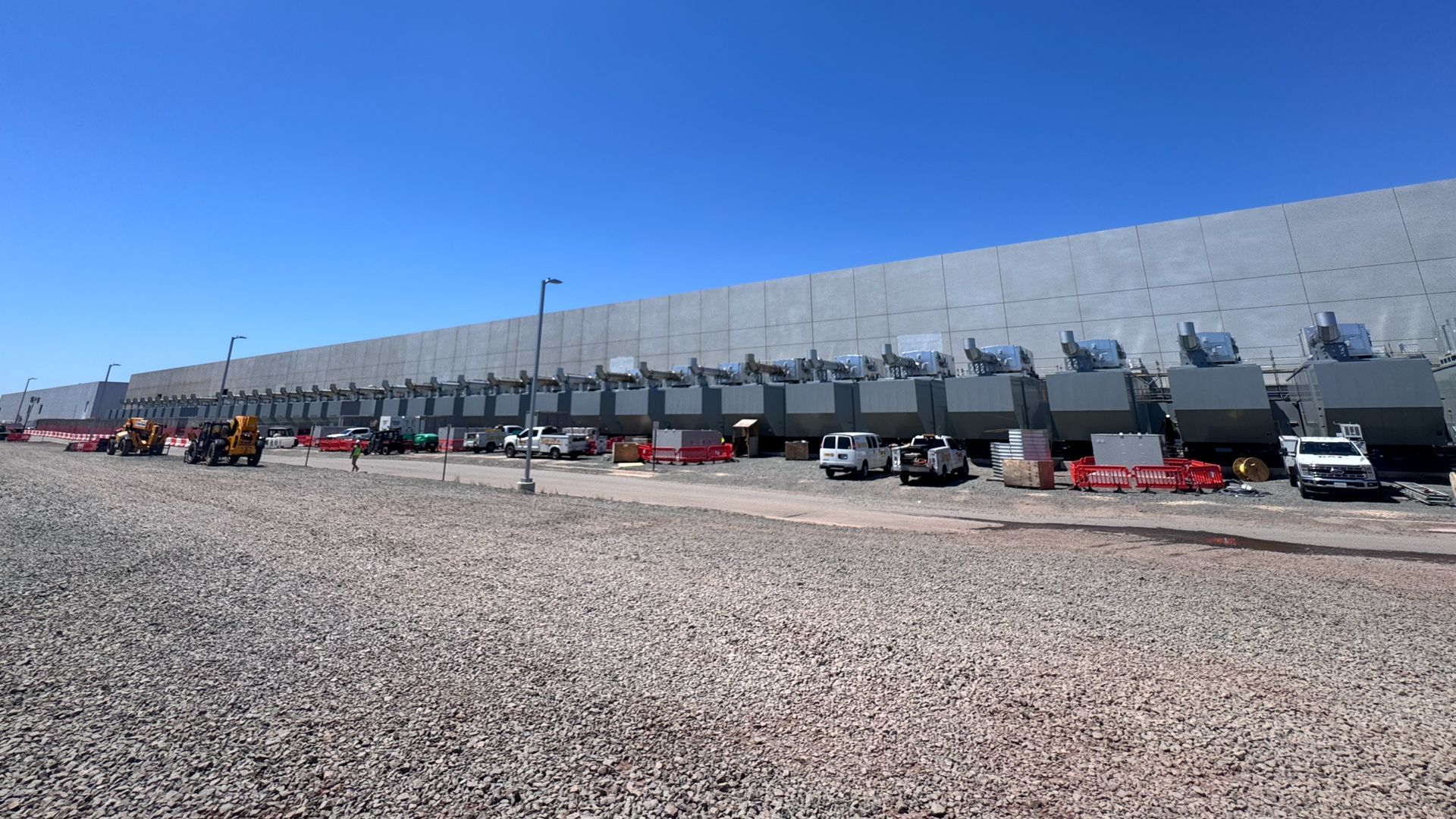 Long gray industrial building with row of large metal ventilation units along base, blue sky overhead; gravel foreground with construction vehicles, red barriers, and parked vans.