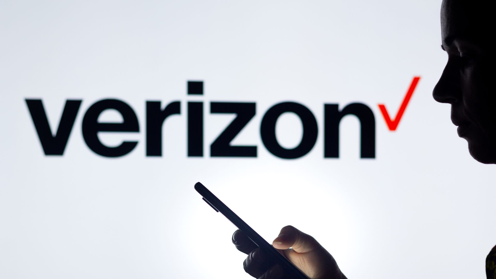 The silhouette of a hand holding a mobile phone in front of the Verizon logo. 