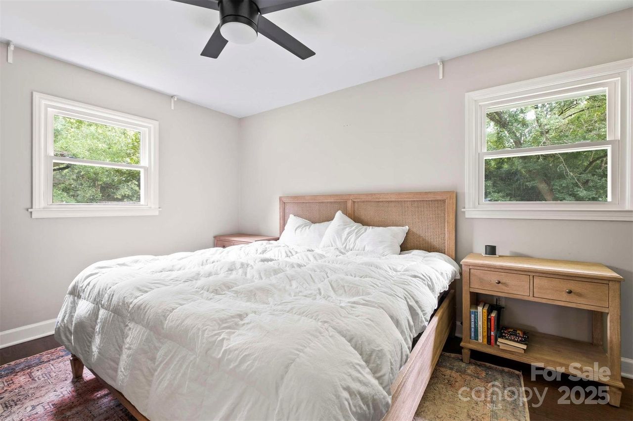 Bright bedroom with white walls, large wooden bed with white bedding, two windows showing green trees, wooden nightstand with books, black ceiling fan.