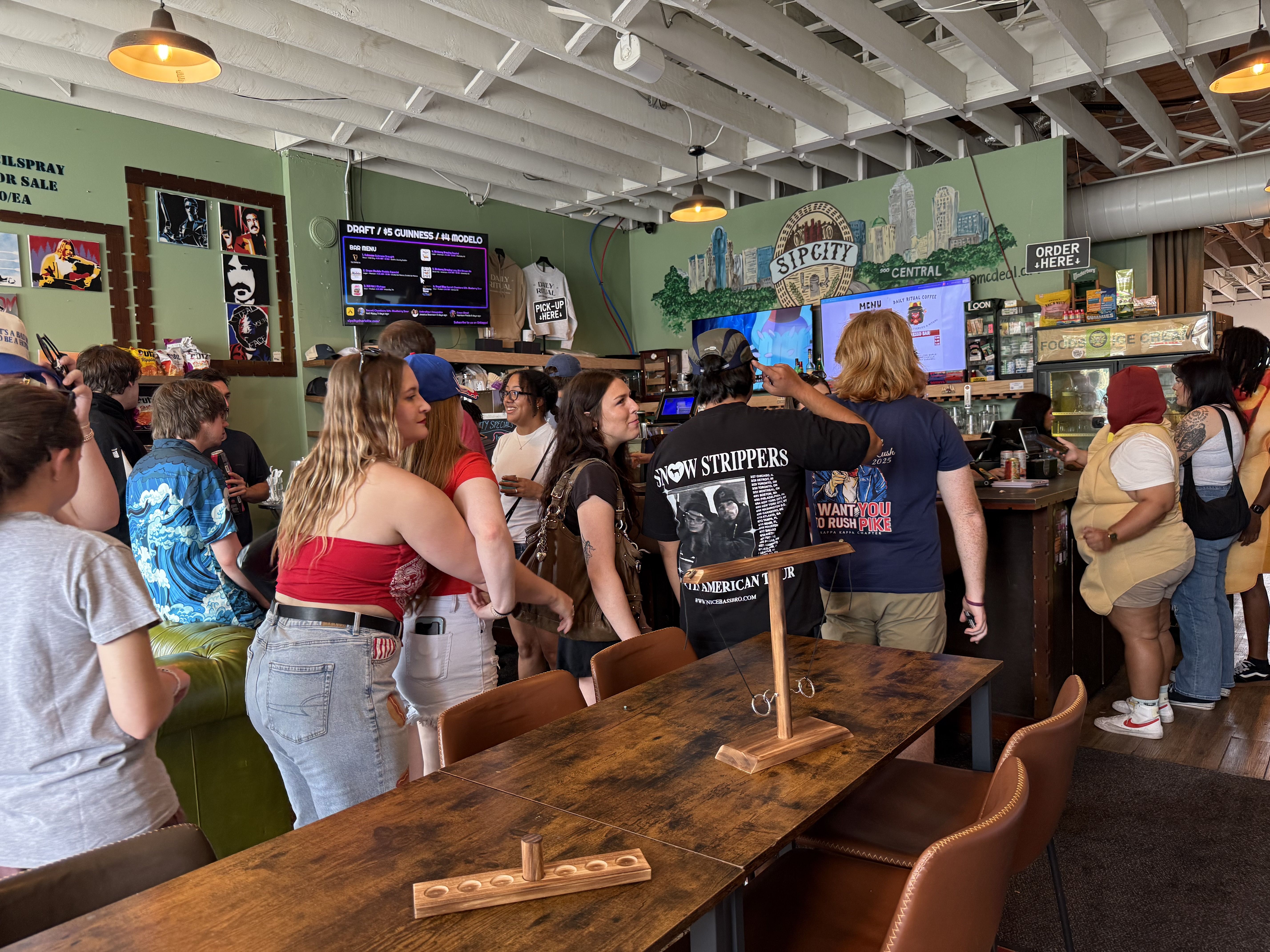 Crowded cafe/bar interior with green walls, a mural Sip City Central, yellow hanging lamps, and a busy counter; people chat around a long wooden table in the foreground.