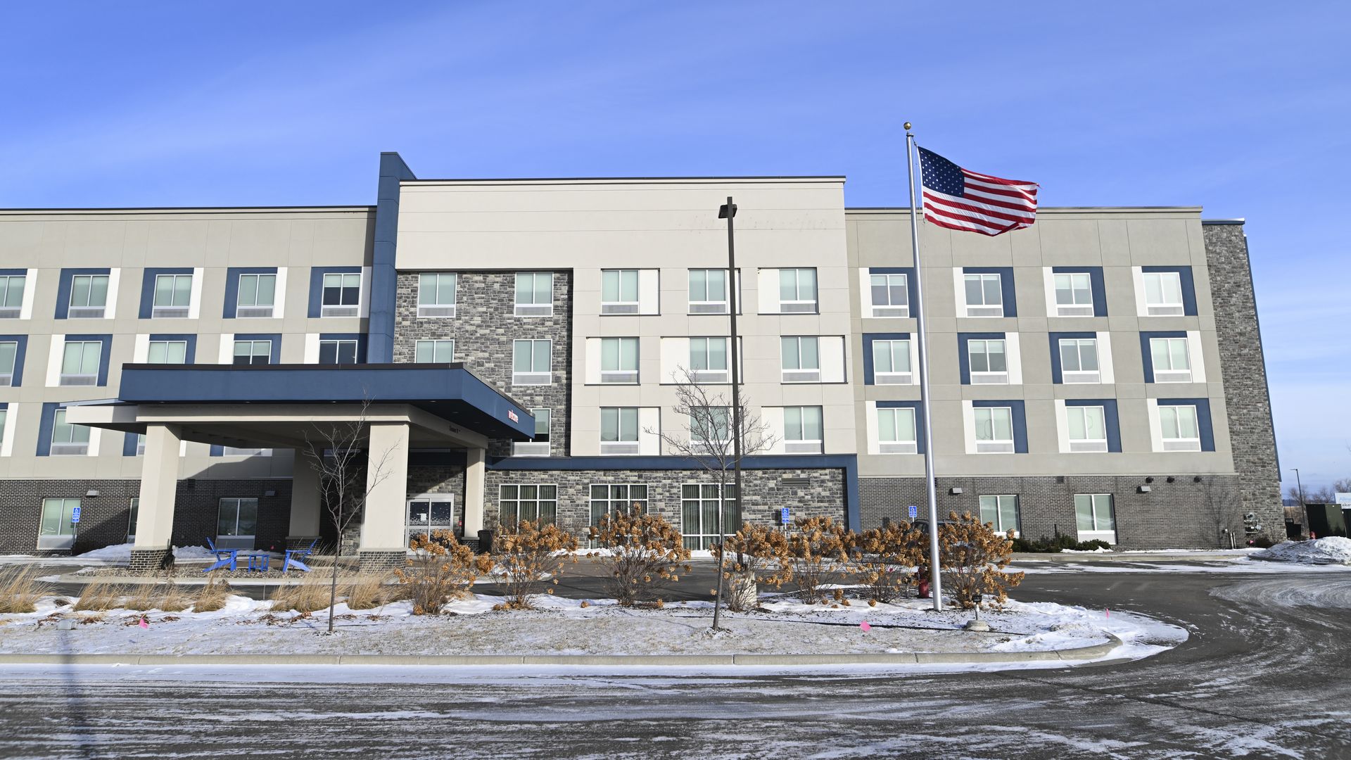 Wide view of a modern, four-story hotel with gray-beige facade and stone accents. A blue entrance canopy, American flag on a tall pole, leafless trees, snow, and a clear blue sky.