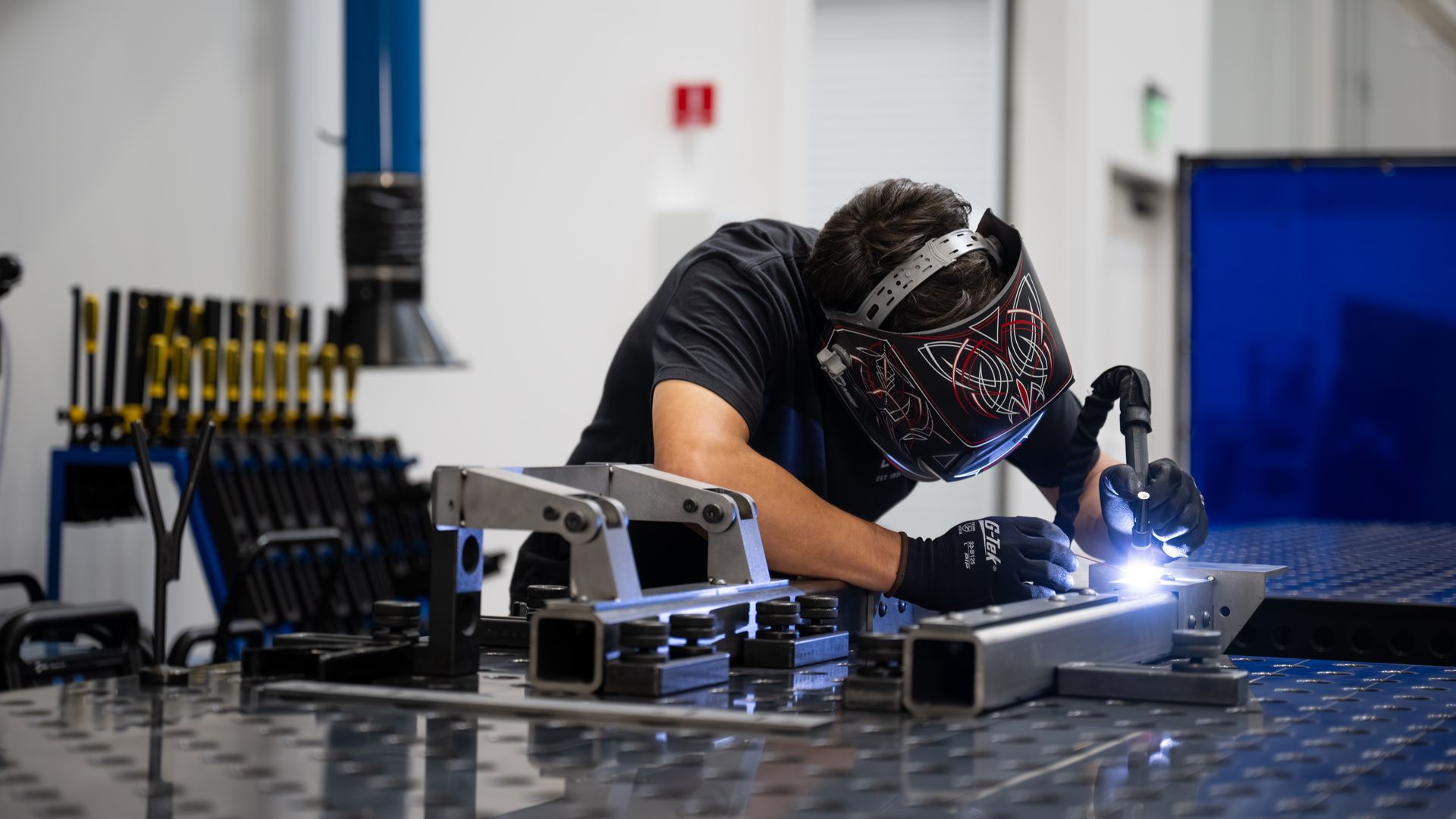 Welder in a patterned helmet and gloves welds a metal frame at a workbench, sparks flying in a bright industrial shop; blue pillar and tool racks visible in the background.