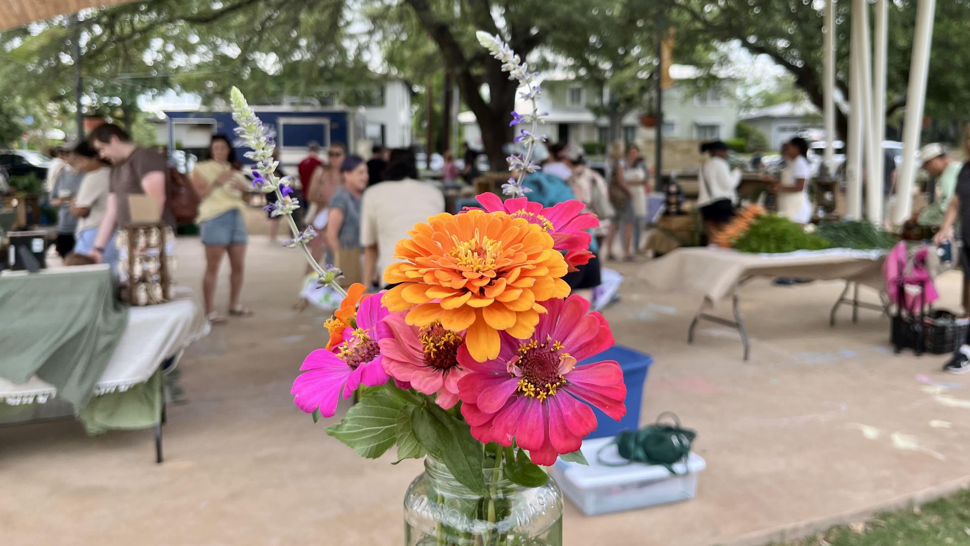 A close-up of a flower bouquet of orange and pink flowers with purple stems in a glass mason jar, set outdoors at a crowded Sunset Ridge Farmers Market under a wooden canopy with blurred people in the background.