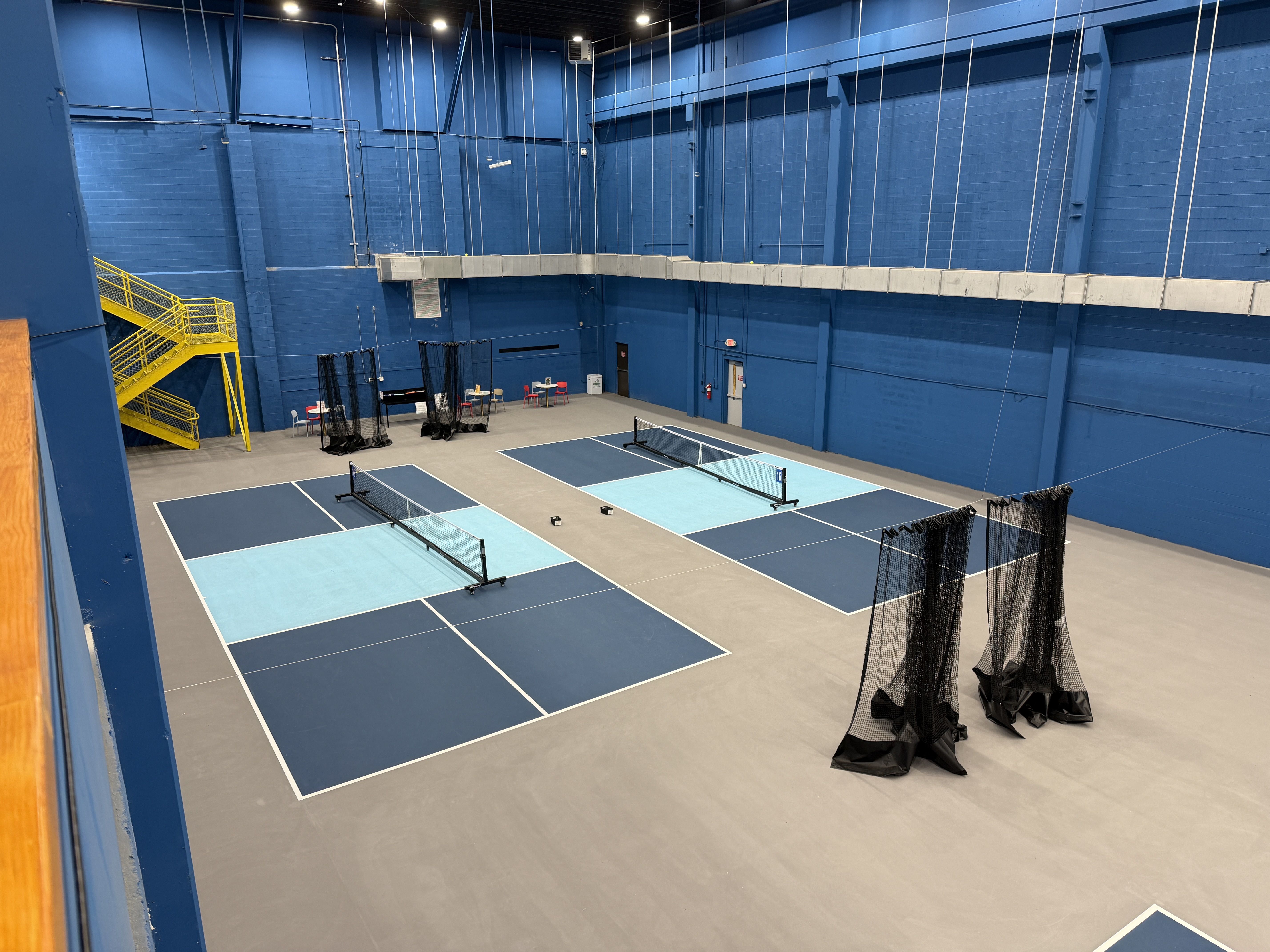 Indoor blue-walled sports hall with several court nets and white boundary lines. A yellow metal staircase on the left, seating area and dividers at the far end.
