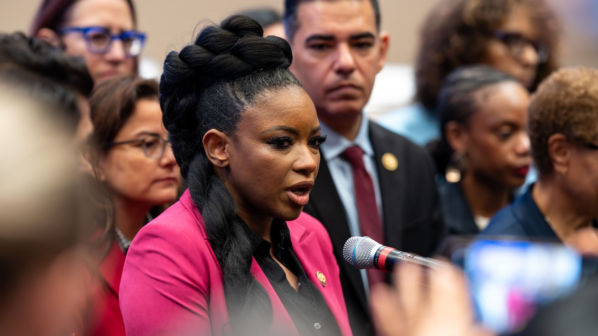 A woman in a bright pink blazer speaks into a microphone at a press event, surrounded by people, some wearing glasses, in a neutral indoor setting.