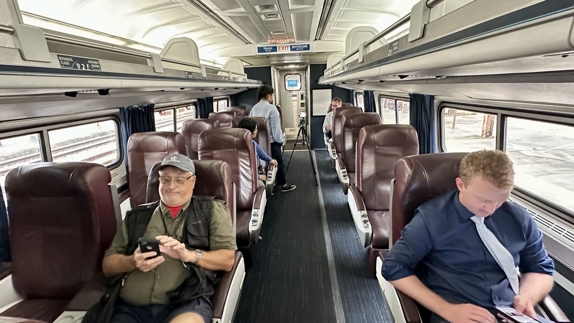 Inside a train's business class carriage with brown leather seats; five passengers engaged with phones or talking; overhead signs for trash, exit, and restroom.