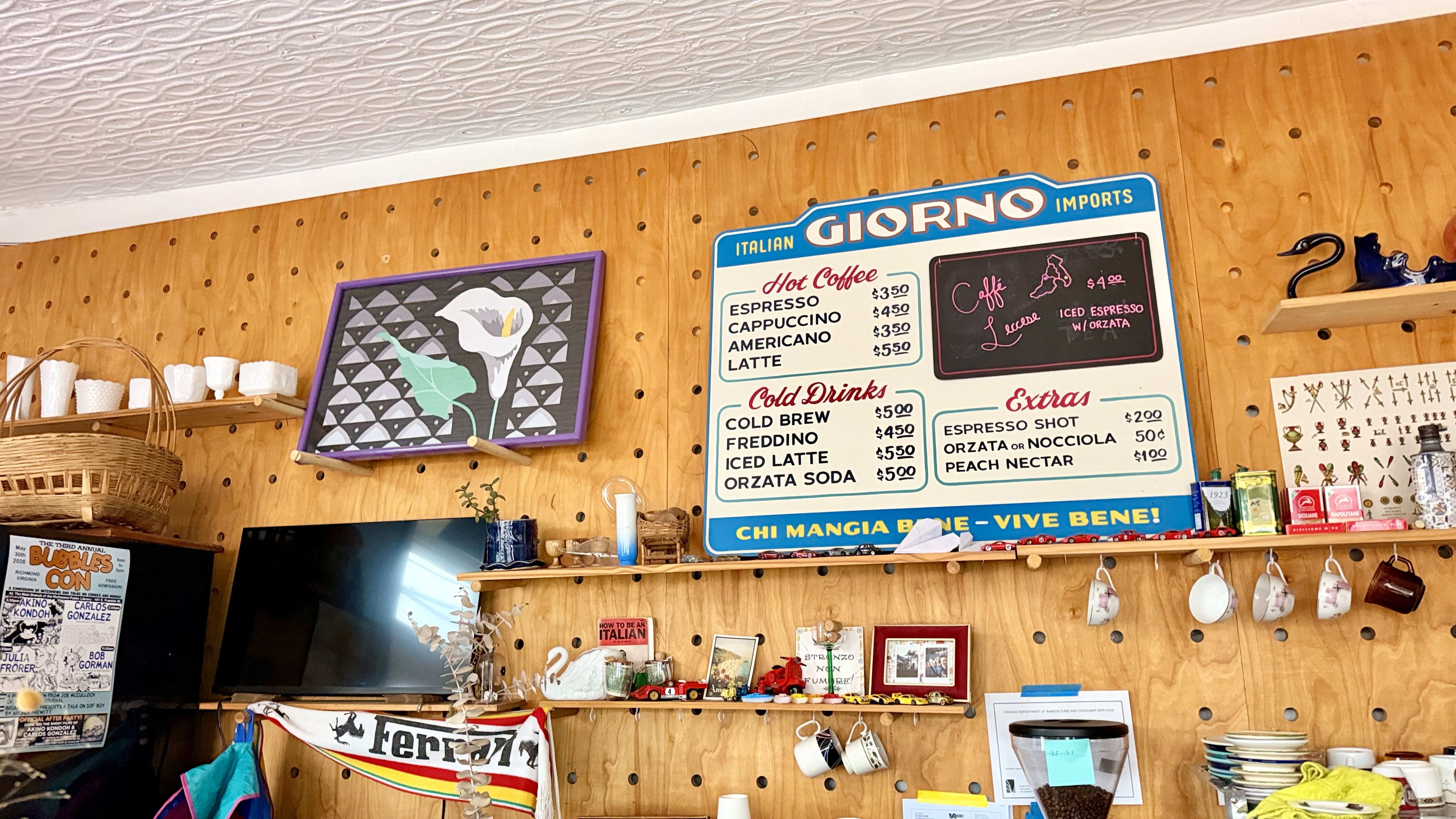 Interior cafe with a pegboard wall, a large blue and white "GIORNO Imports" menu board, purple-framed art, wicker basket, and shelves of mugs and trinkets.