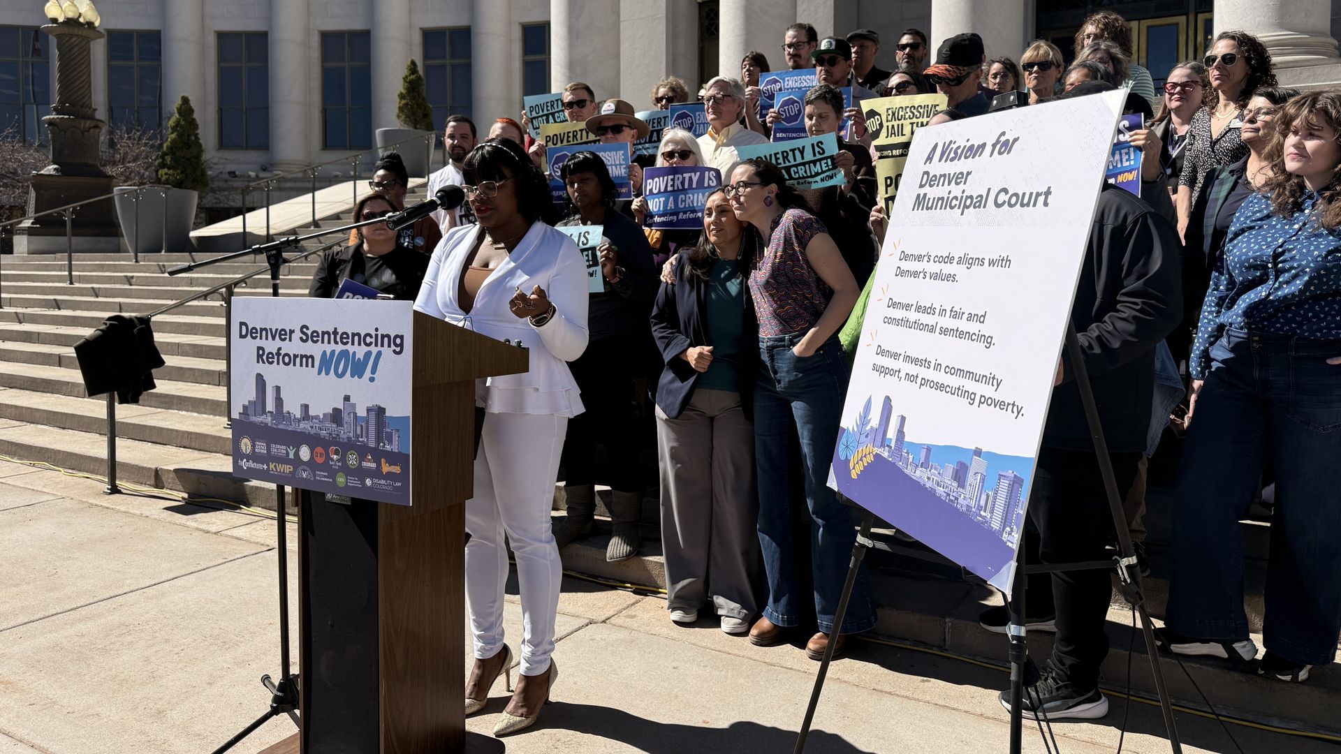 Speaker in white at courthouse steps, addressing protesters holding signs like Denver Sentencing Reform NOW! and a large board reading A Vision for Denver Municipal Court.