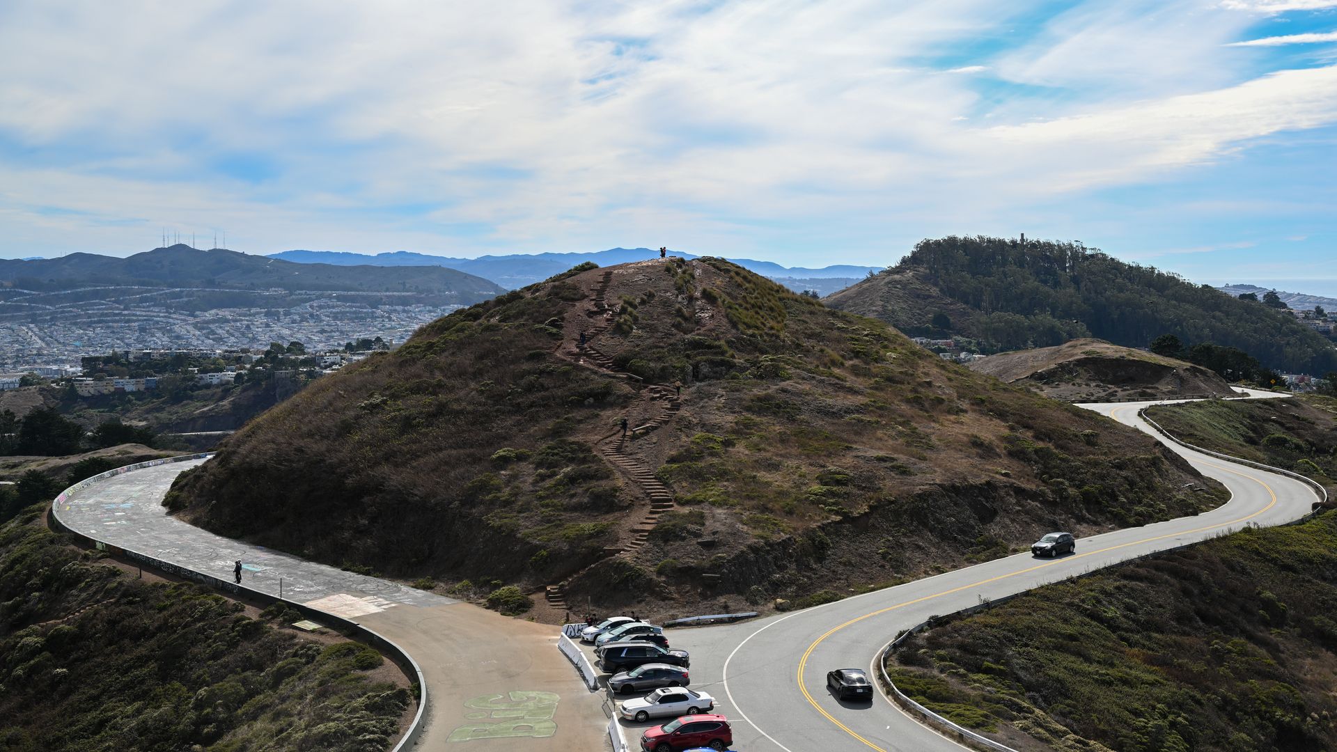 Brown hills with a winding stair path to a hilltop where a person stands. A curved road with parked cars circles the slope, while a distant city and hills lie under a blue, cloudy sky.