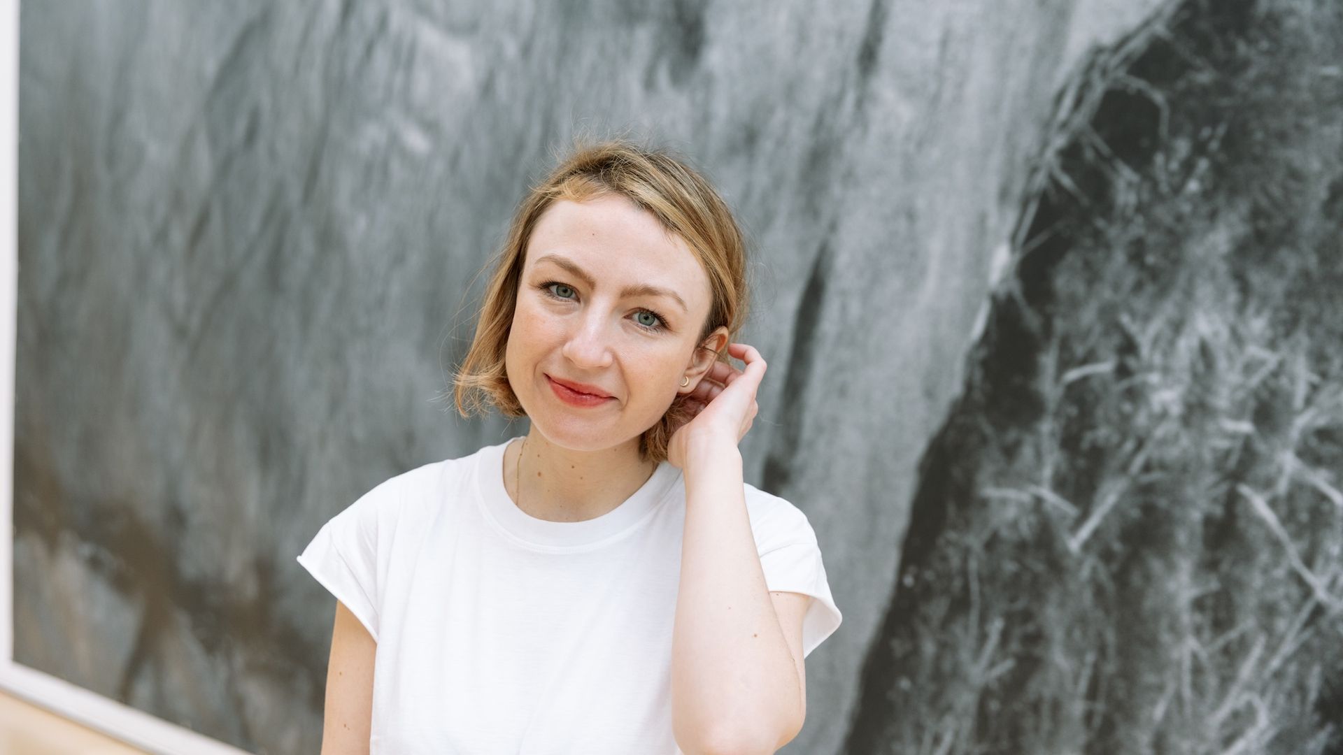 Rowan Beaird smiling with short blonde hair wearing a white T-shirt stands before a large gray marble-like backdrop, tilting her head and touching her ear.