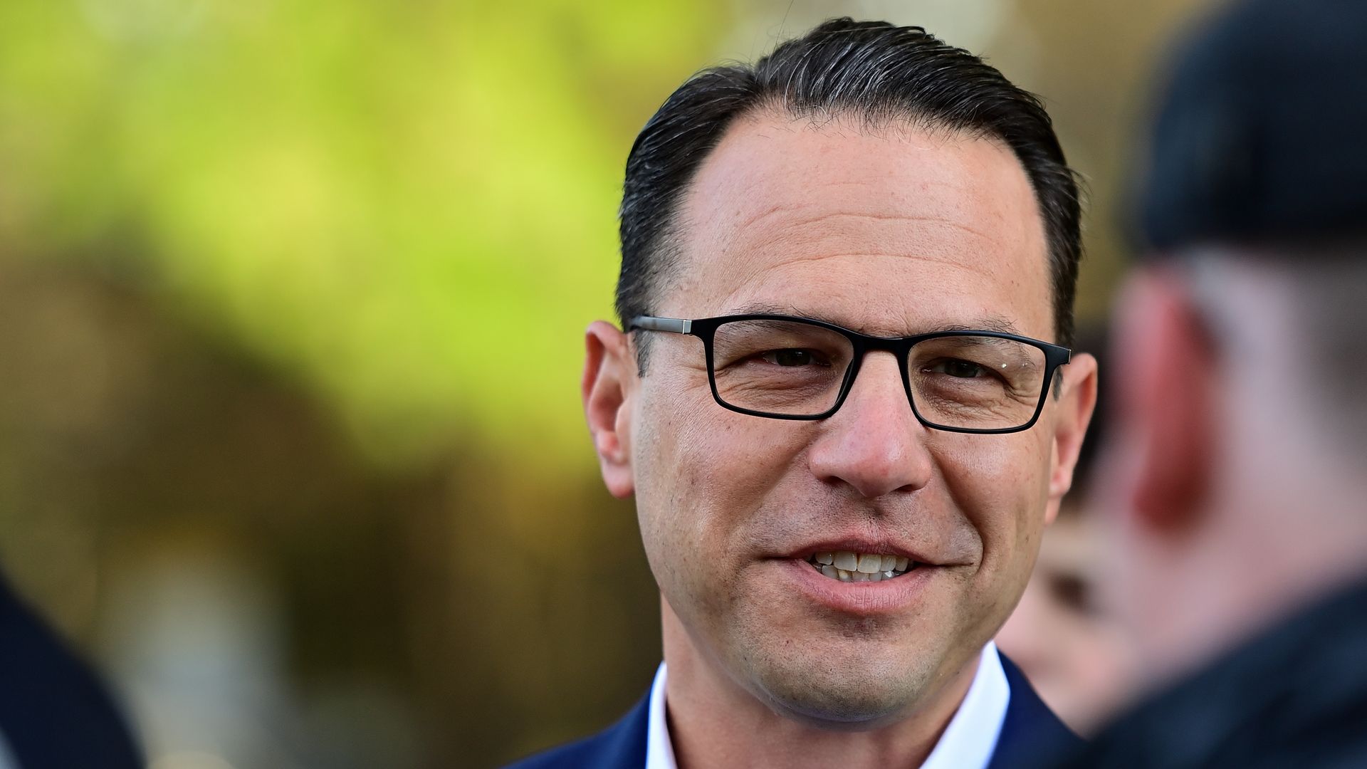 A close up of a man's face who has short, dark hair and is wearing black, rectangular glasses. 