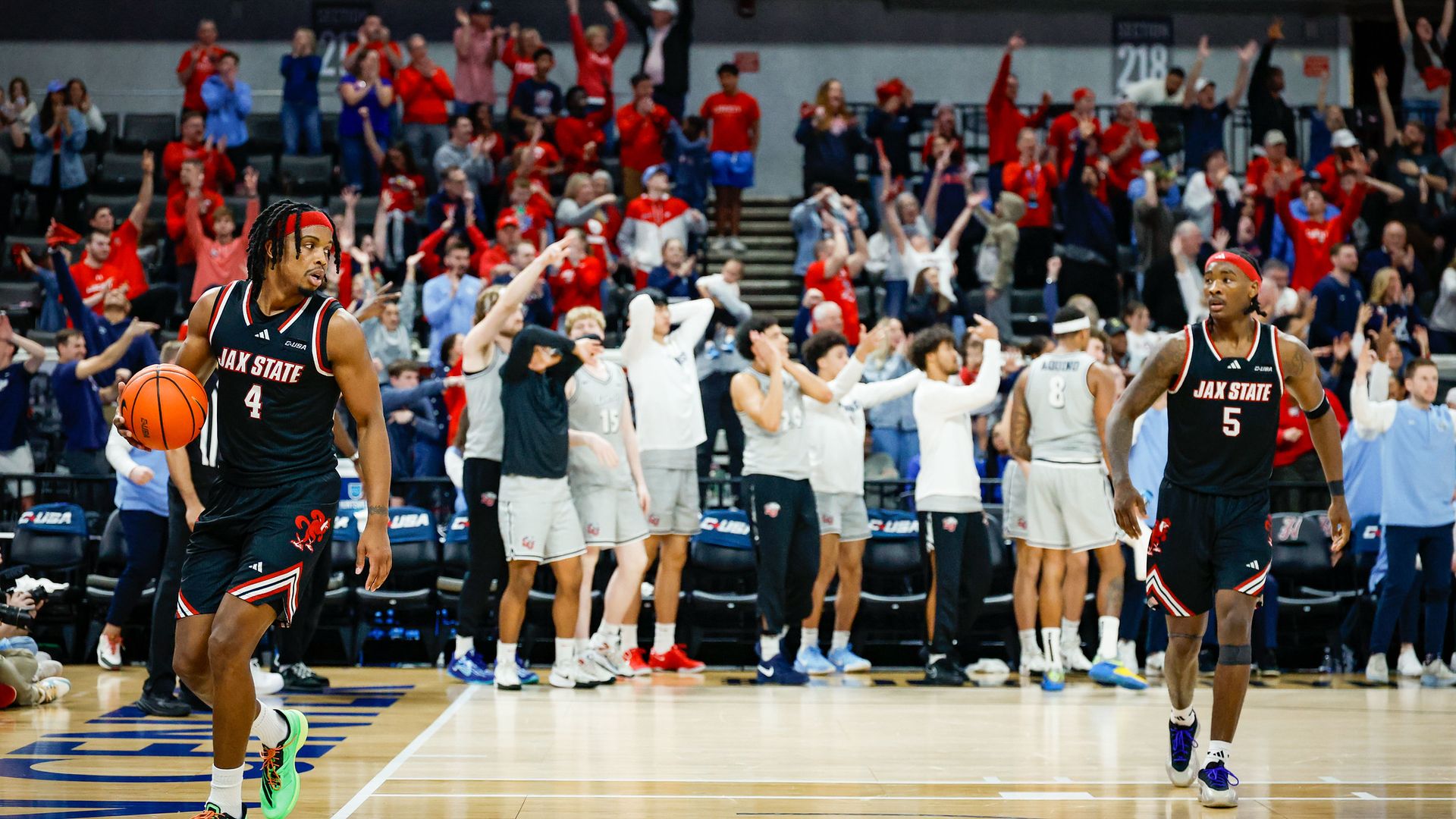 Two basketball players from JAX STATE in black uniforms, one holding a basketball, on court. Behind them, a crowd and opposing team in gray react with mixed emotions during a game.