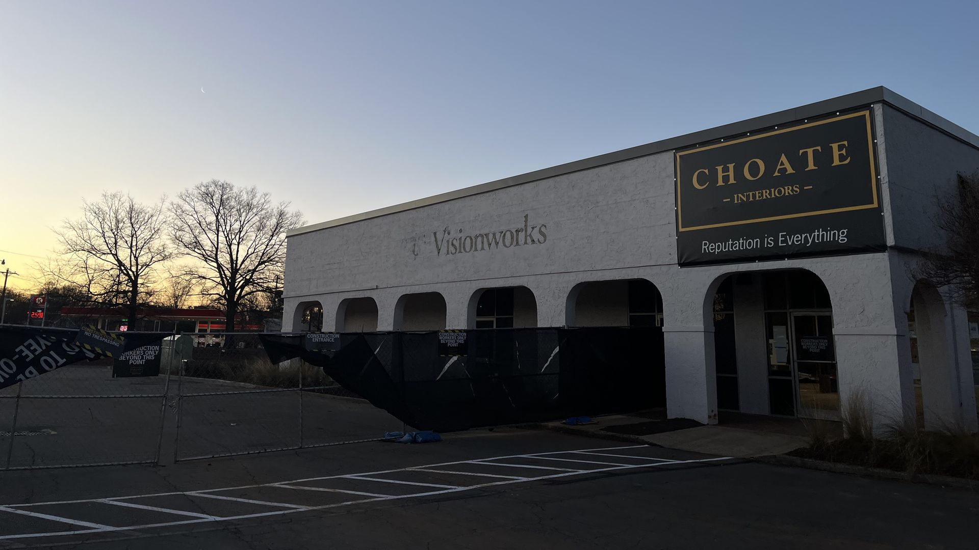 Sunset sky over a white commercial building with signs for Visionworks and Choate Interiors, fenced off for construction with black barriers and bare trees in the background.